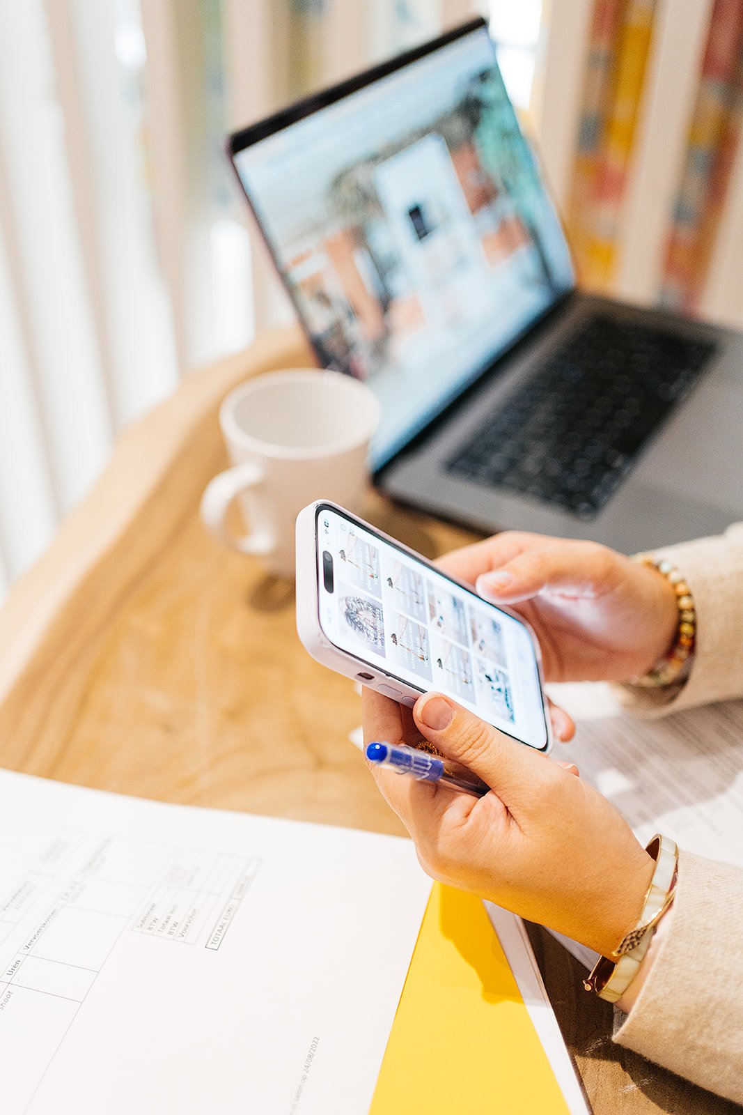 Person holds a smartphone with photo or gallery page in hand, laptop and a white mug on a wooden table, in a bright room.