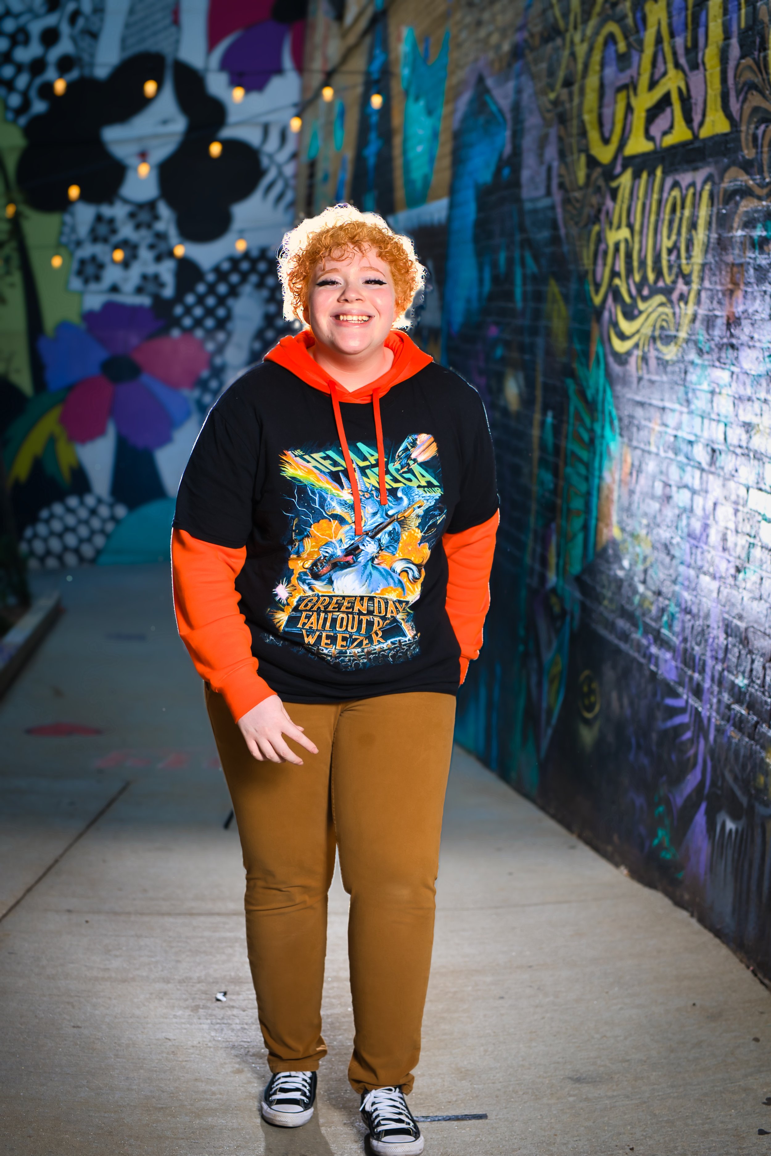 Person smiling in colorful alley with murals, wearing a concert-themed hoodie and casual pants.