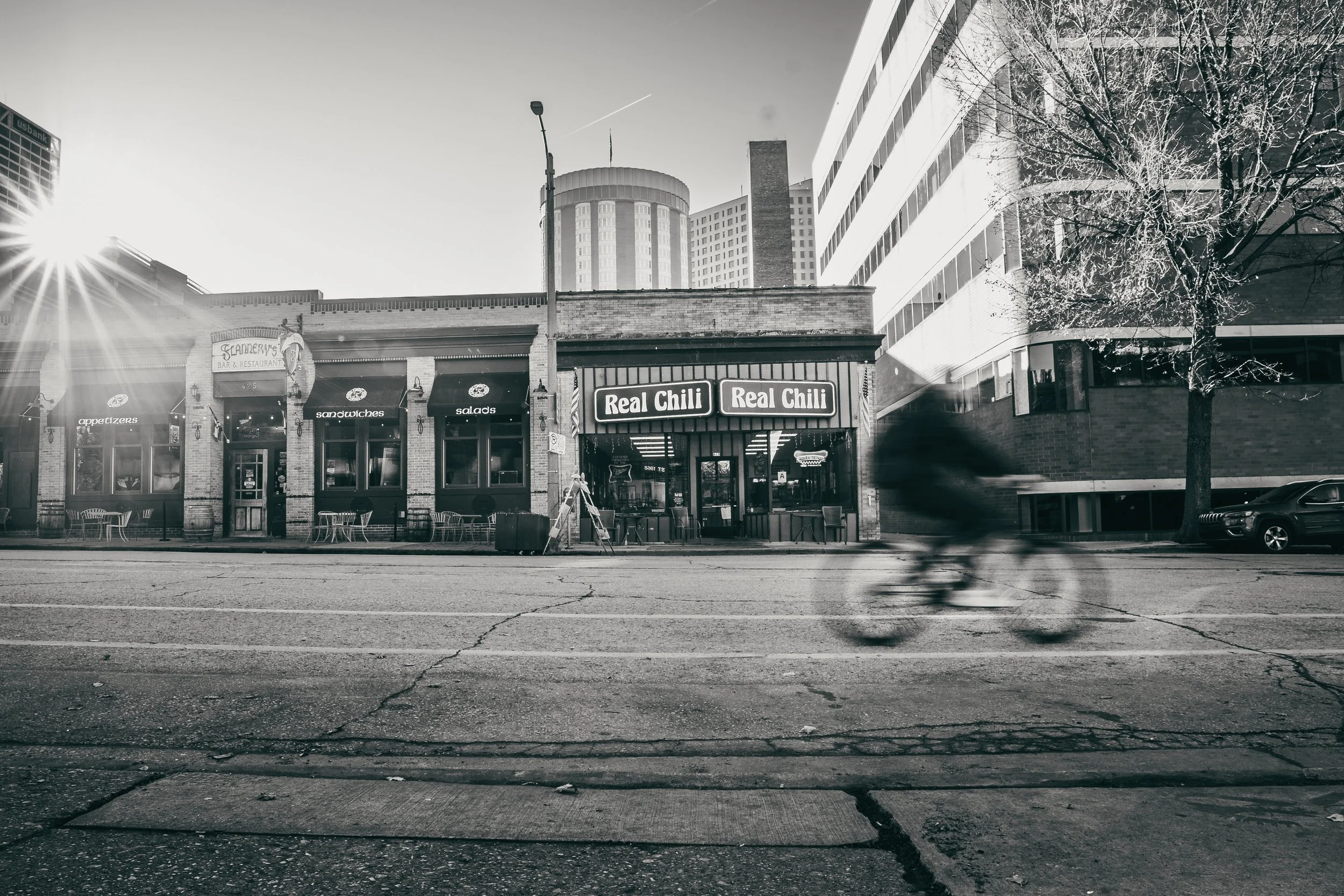 A city street with a bar named 'Stanfrey's Bar & Restaurant' and a restaurant called 'Real Chili' in a mixed urban environment, blurred cyclist riding past, trees, cars, and tall buildings in the background, black and white photo.