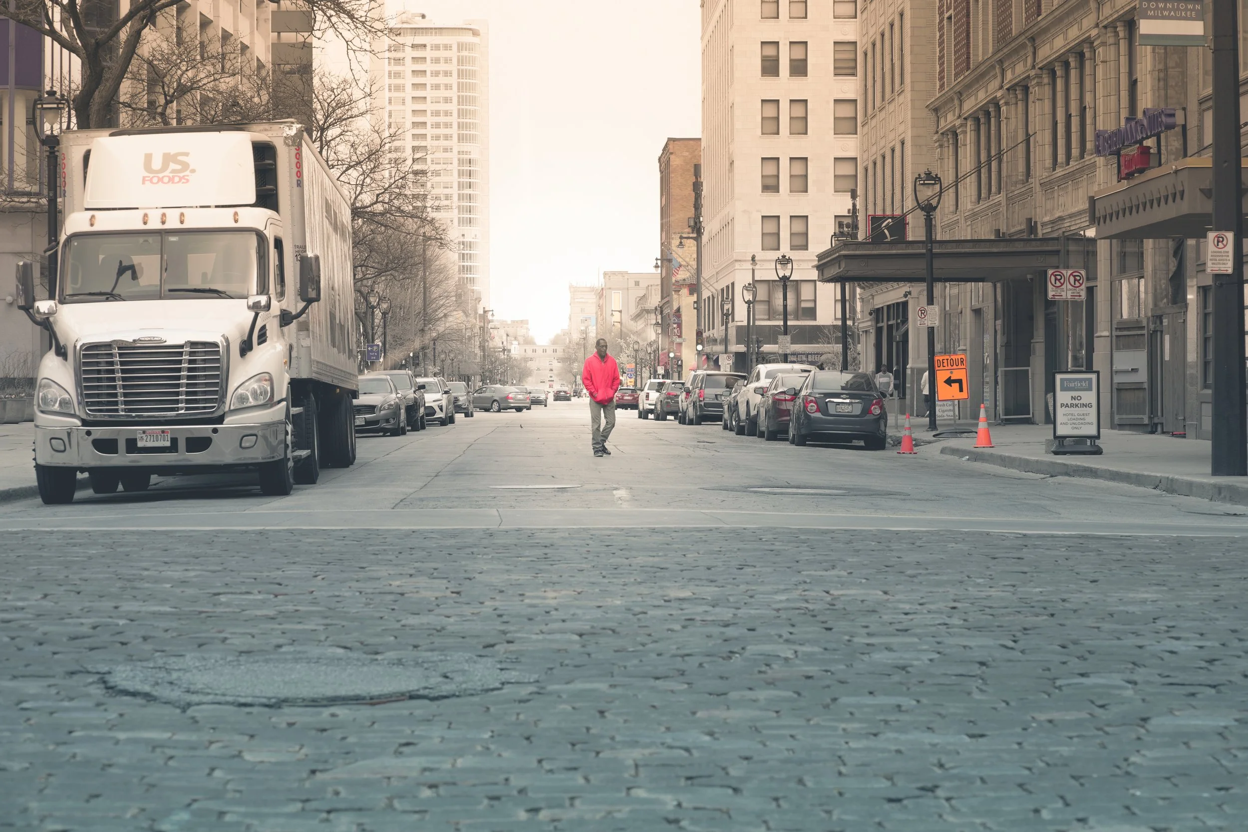 A city street scene with a white delivery truck and parked cars lining the curb. A person wearing a bright pink jacket walks in the middle of the street. Tall buildings with storefronts and signs are visible on the right, with street signs and orange