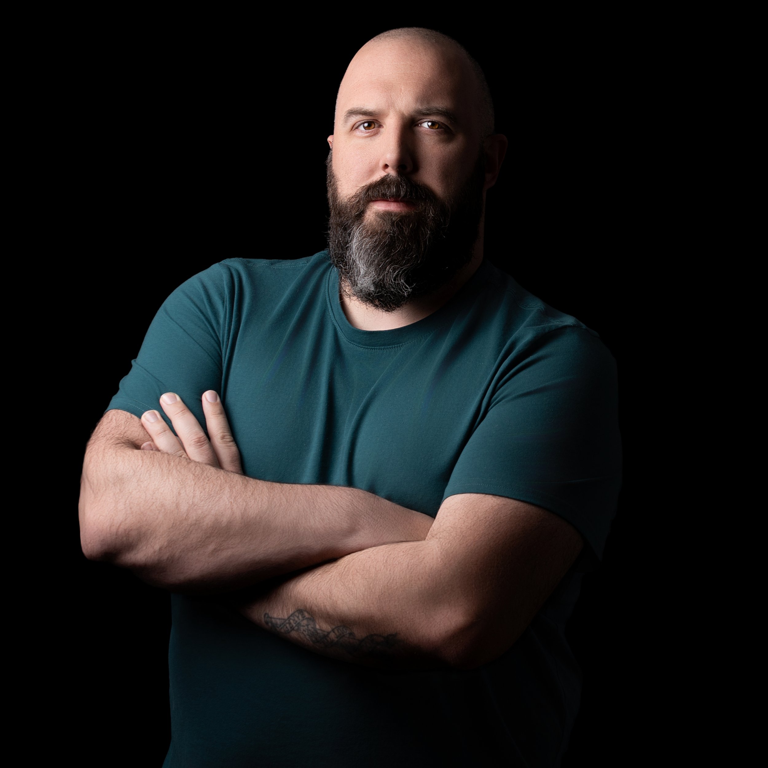 Bald man with a beard in a teal shirt standing with arms crossed against a black background.