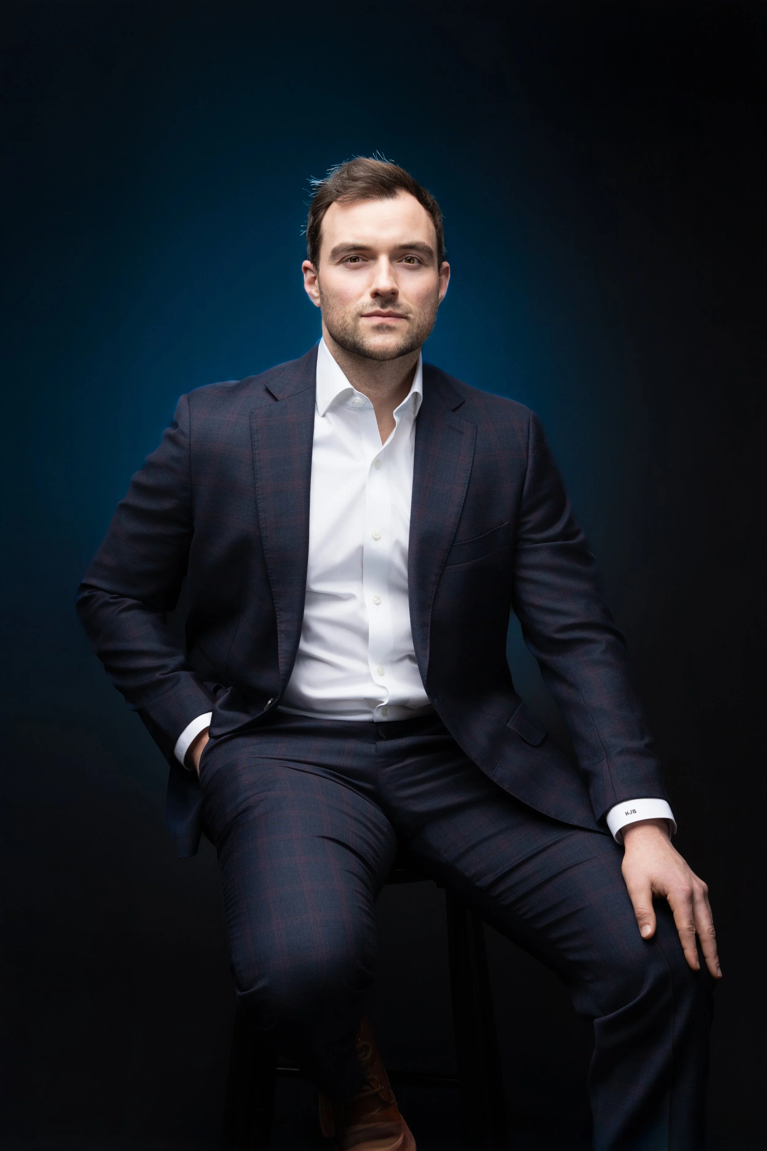 A man in a dark plaid suit and white shirt sitting on a stool against a dark background.