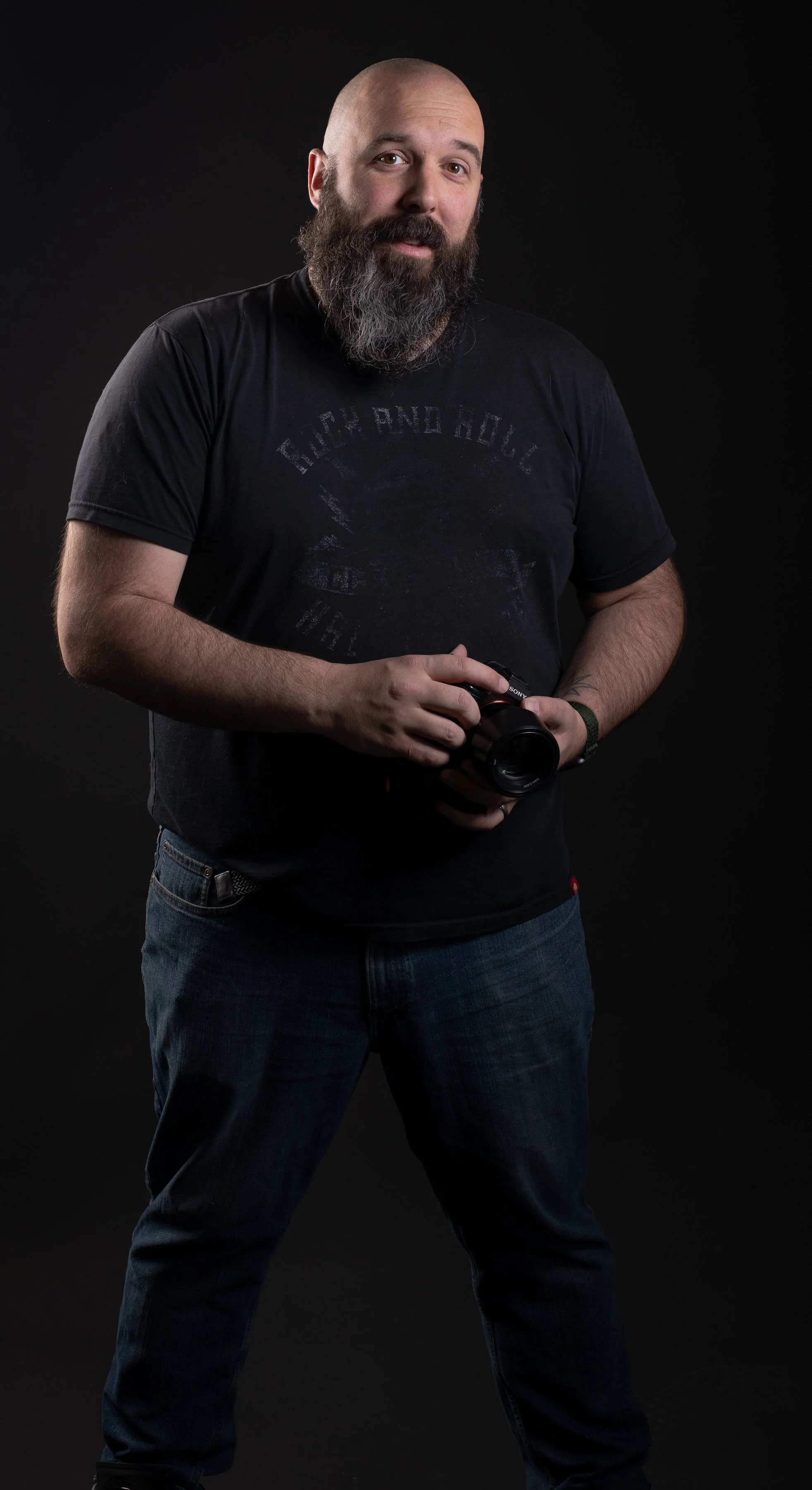 A man with a beard and shaved head holding a camera, standing against a black background.