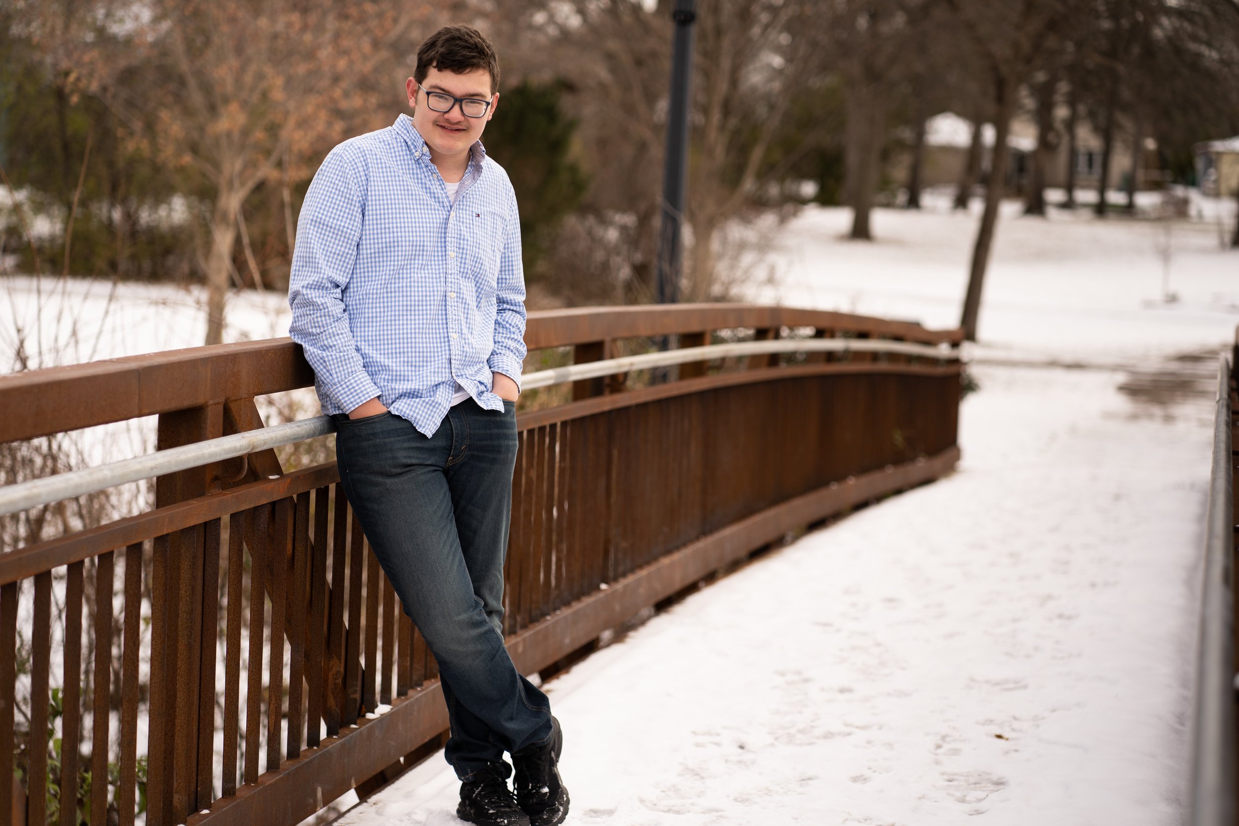 Person in glasses and checked shirt leaning on a bridge railing in a snowy park.
