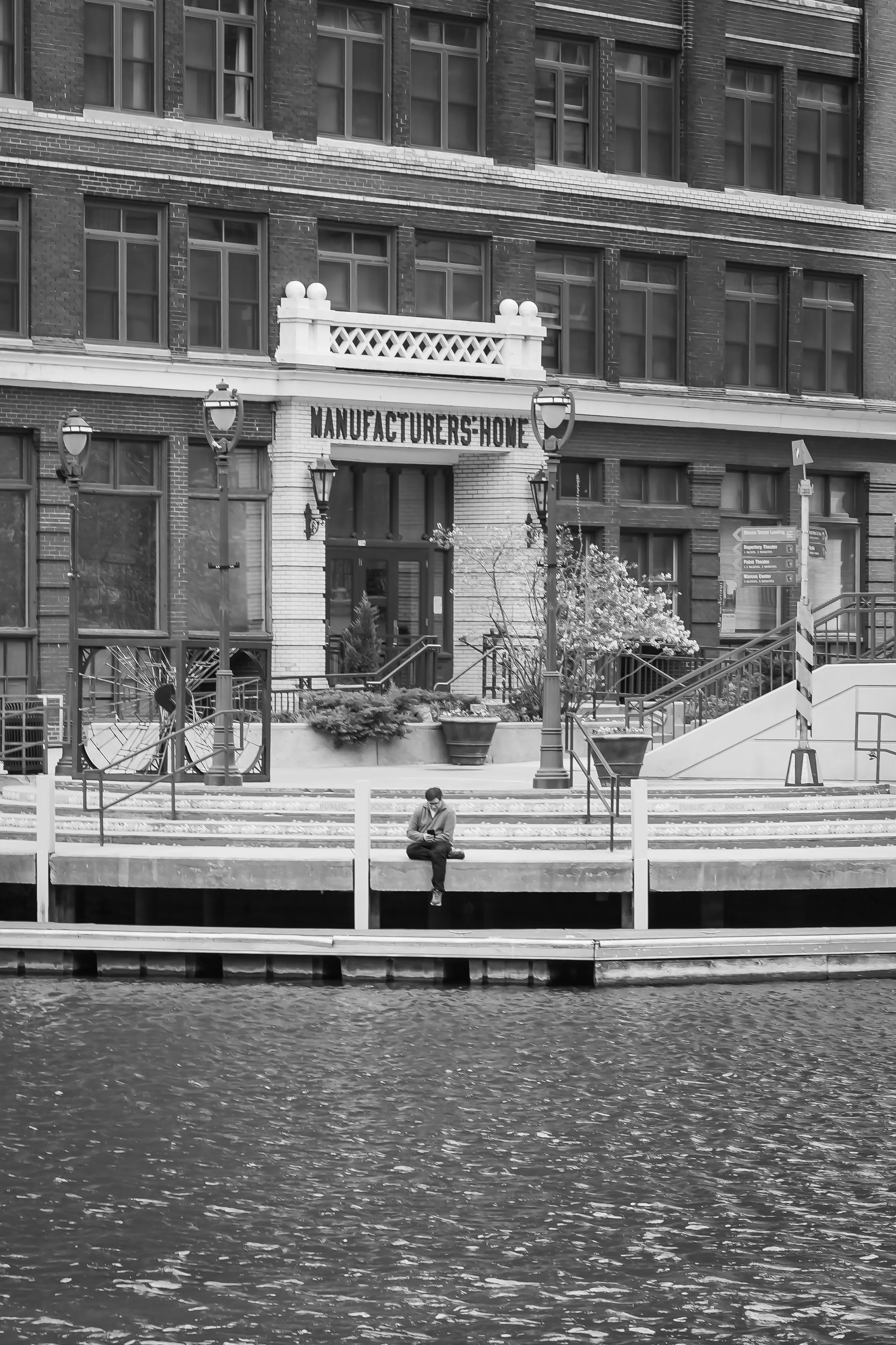 A man sitting on a dock by water in front of a brick building with white accents, street lamps, planters, stairs, and a sign that reads 'Manufacturers Home'.