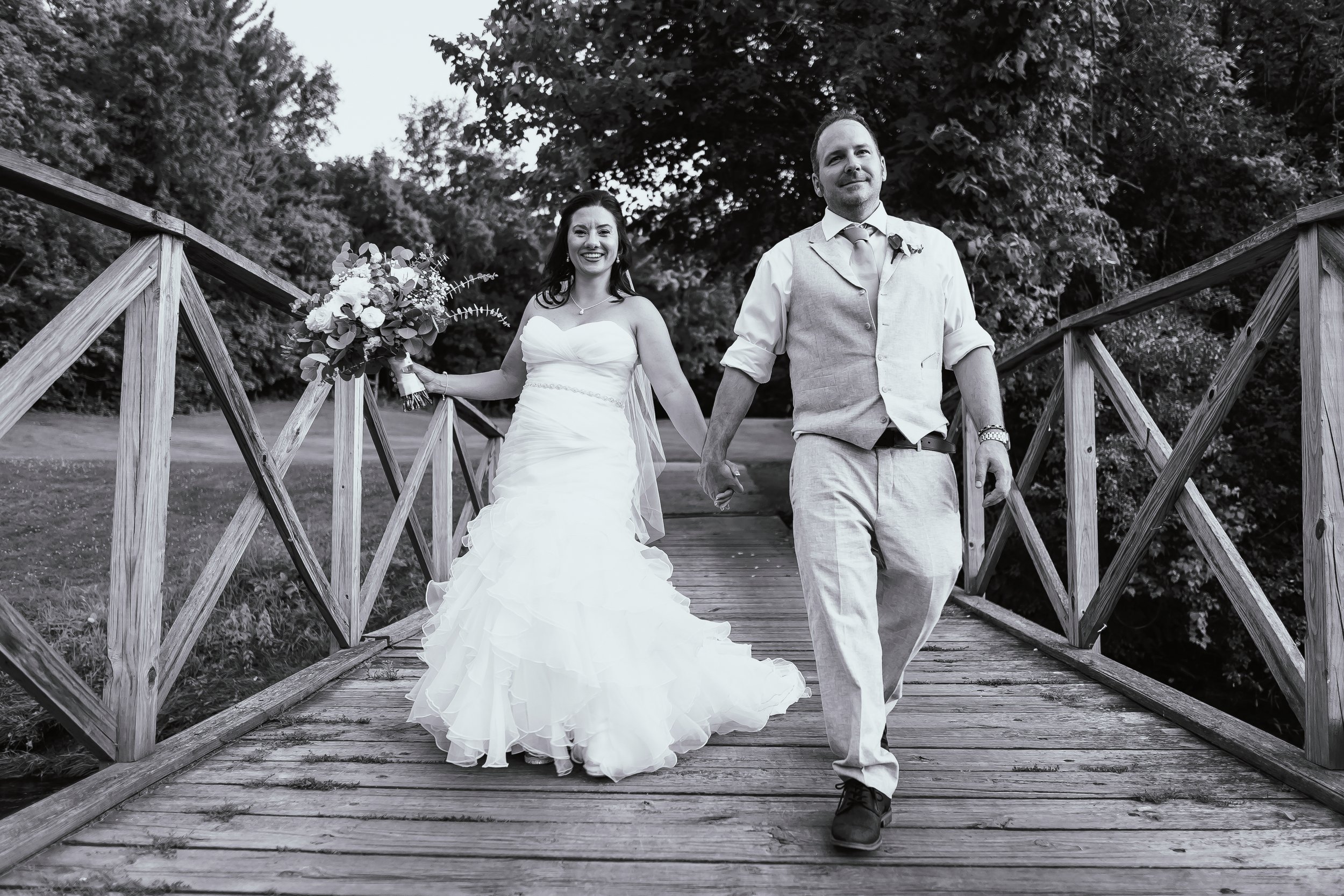 Wedding couple holding hands on a wooden bridge, bride in a strapless gown holding a bouquet, with trees in the background.