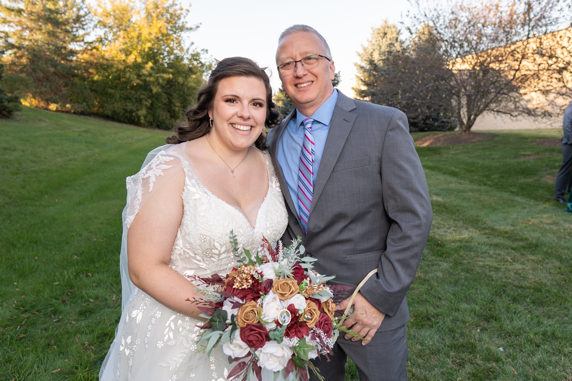 Bride in a white wedding dress holding a colorful bouquet, standing next to a man in a gray suit and striped tie, both smiling, outdoors with greenery in the background.