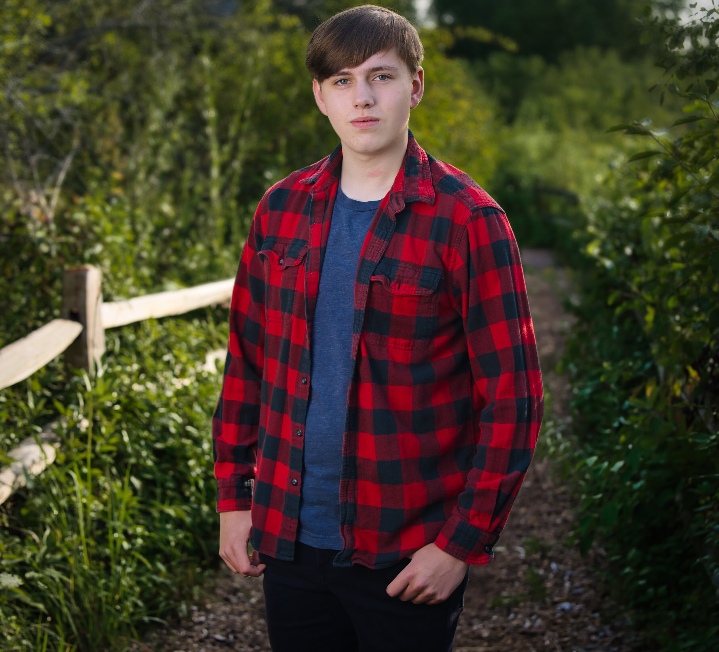 Teenage boy wearing a red and black plaid shirt stands on a path surrounded by greenery.