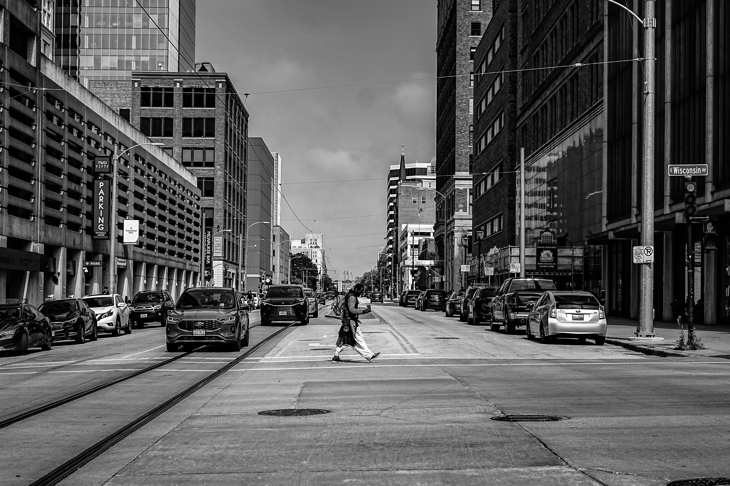 A black and white photo of a city street with parked cars on both sides, high-rise buildings, and a person crossing the street wearing a backpack and a long skirt.