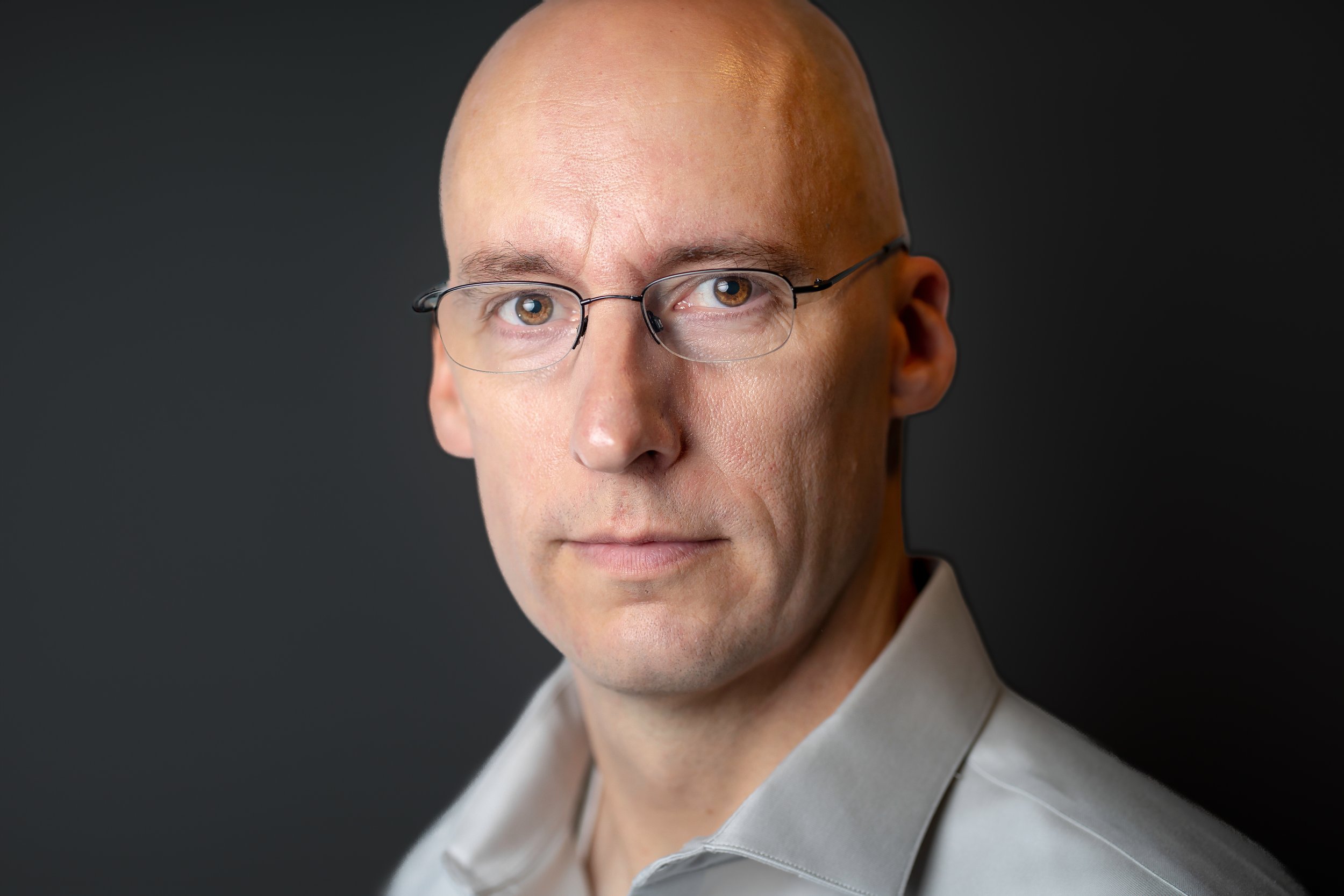 Bald man wearing glasses and a white shirt against a dark background.