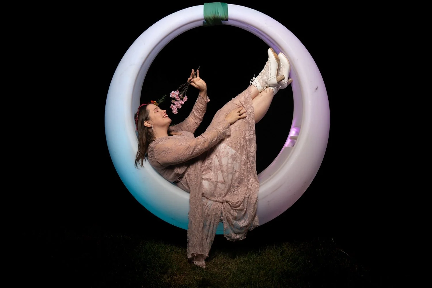 A woman in a lace pink dress and white boots sitting inside a large illuminated white ring, holding a small bouquet of pink flowers, against a black background.