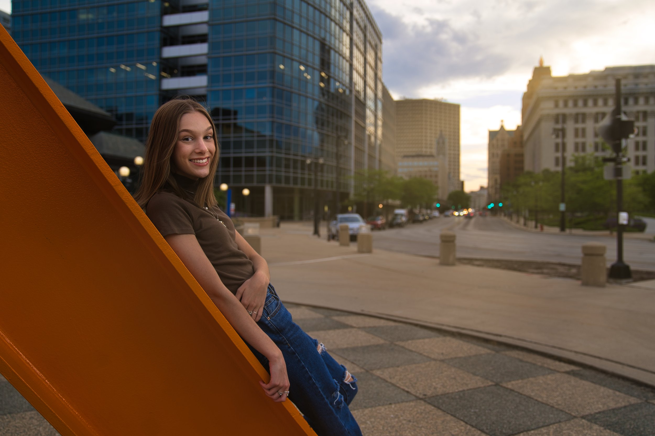 Smiling person leaning on an orange sculpture in an urban setting with tall glass buildings and a sunset in the background.