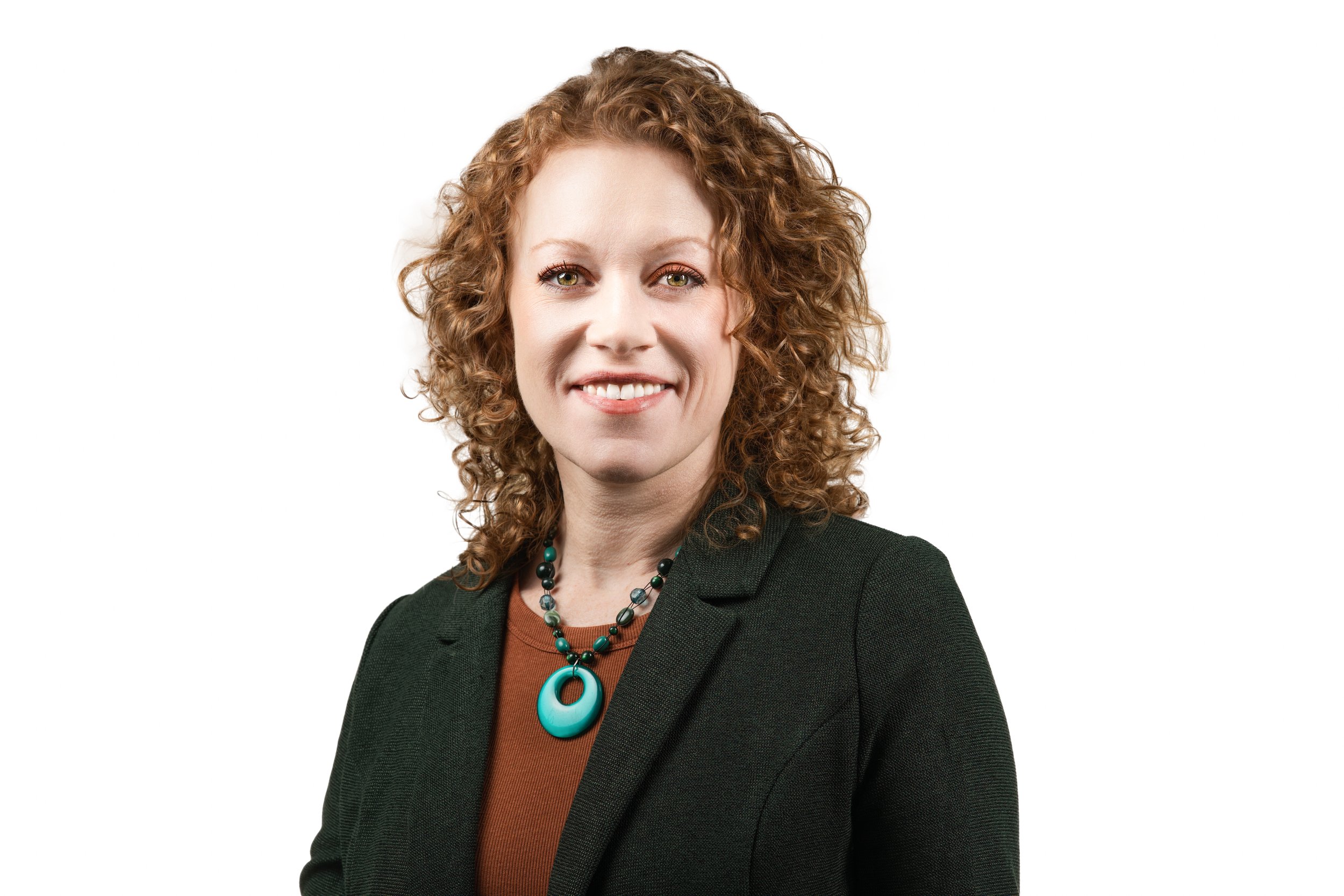 Person with curly hair, wearing a blazer and necklace, smiling against a white background.