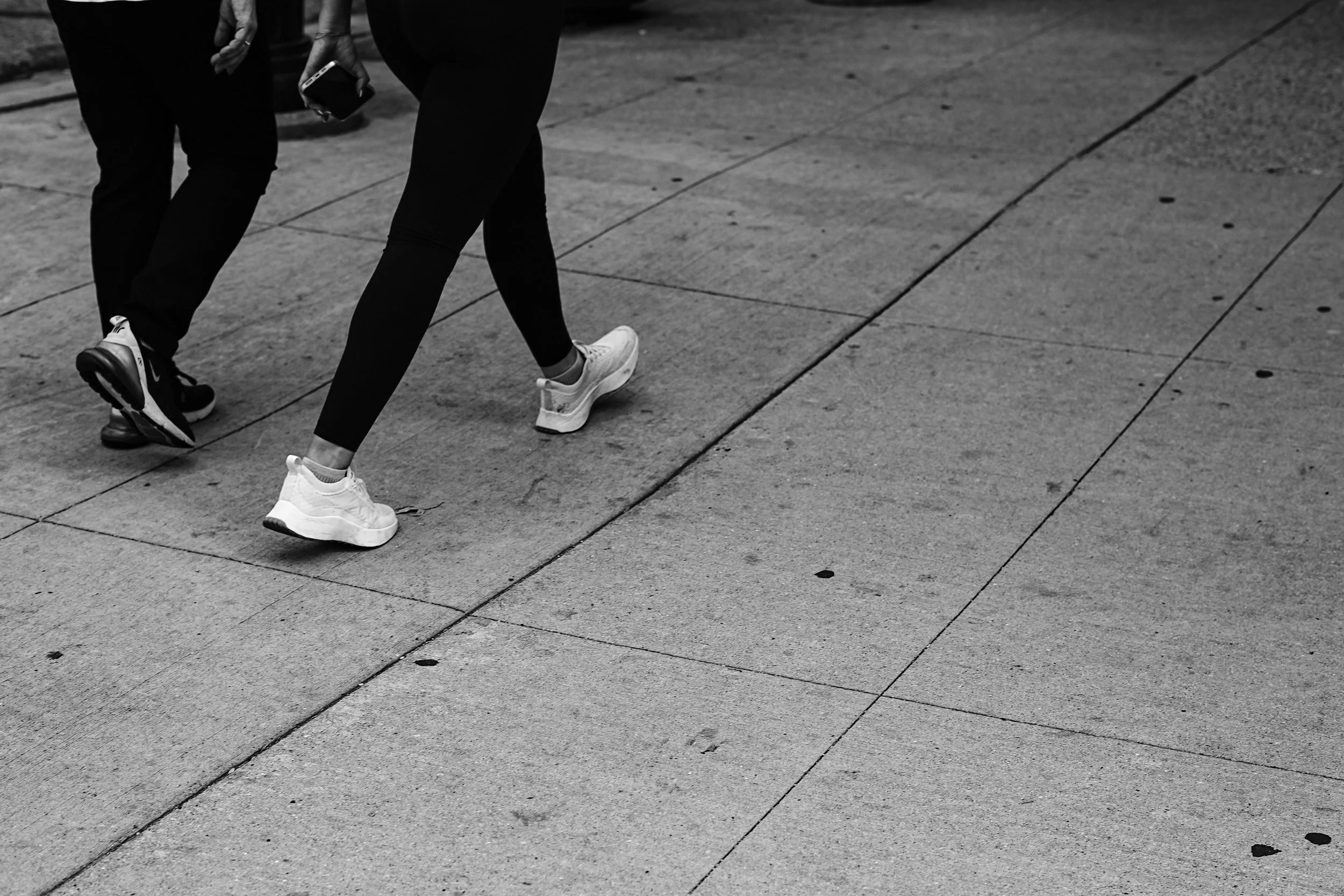 Close-up of the lower bodies of two people walking on a city sidewalk, wearing athletic shoes and black pants.