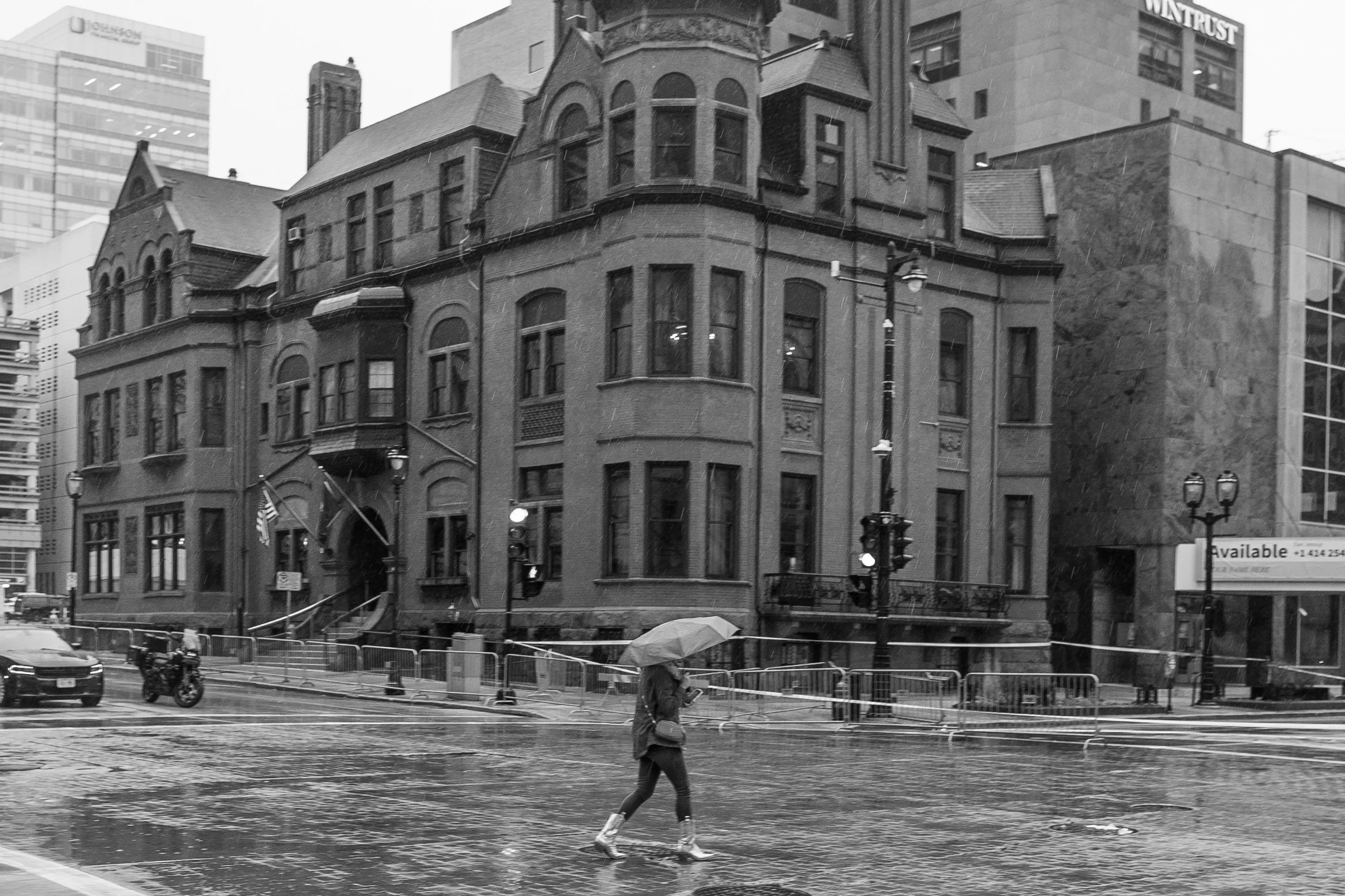 A person crossing the street with an umbrella in rainy weather, in front of old brick and stone buildings in a city.