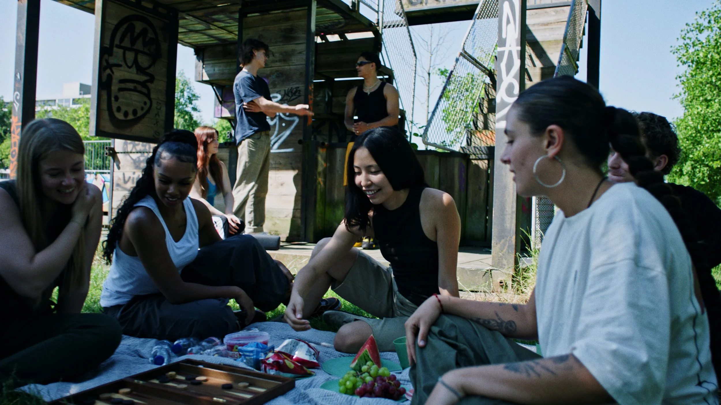 Een groep jonge mensen zit op een mat op het gras, die samen eten en lachen, in een buitenomgeving met een houten structuur en graffiti.