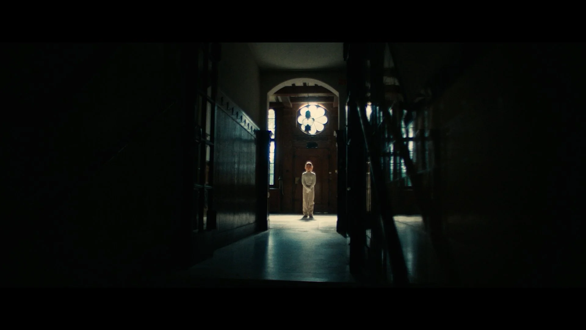 Een persoon staat in een donkere gang met licht dat op hem of haar valt, achter een houten deur en een rozetraam.