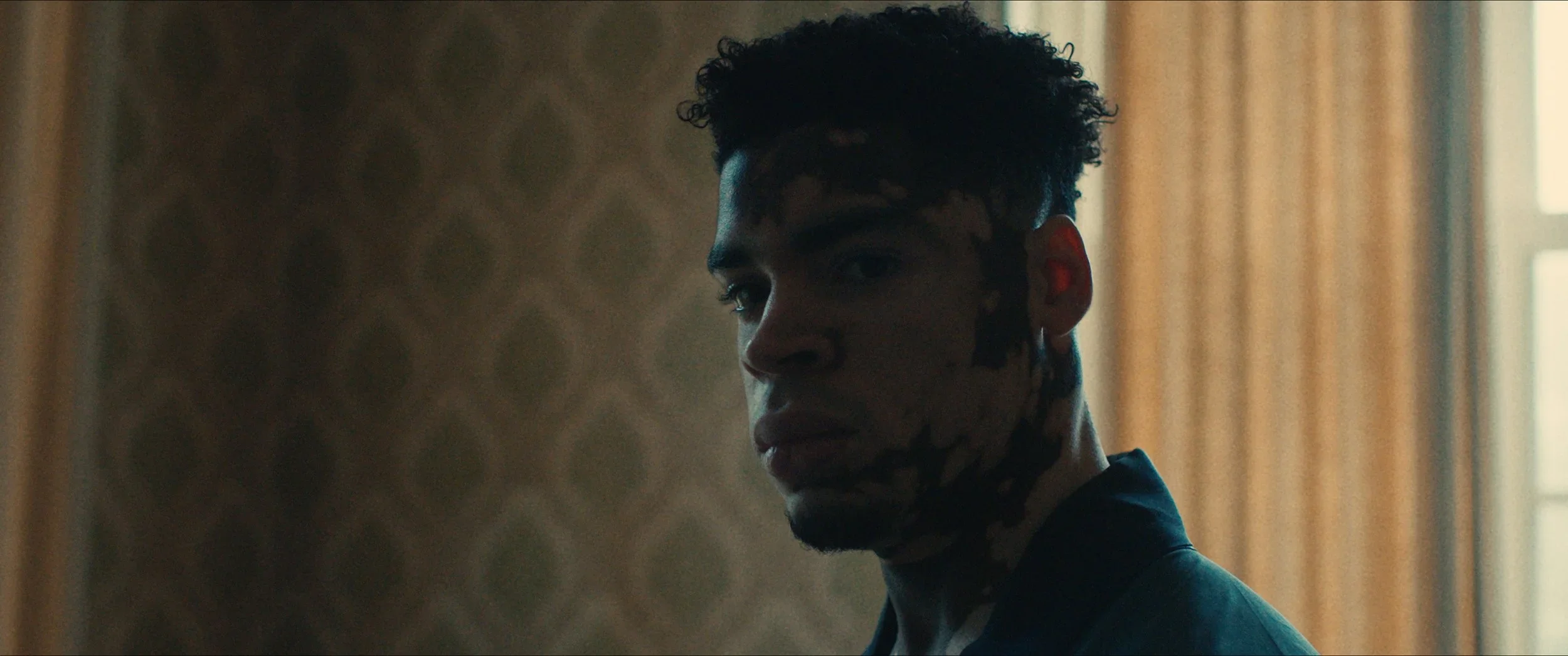 Een jonge man met kort, krullend haar kijkt over zijn schouder naar de camera, met een achtergrond van een kamer met een raam en patronen op de muur.