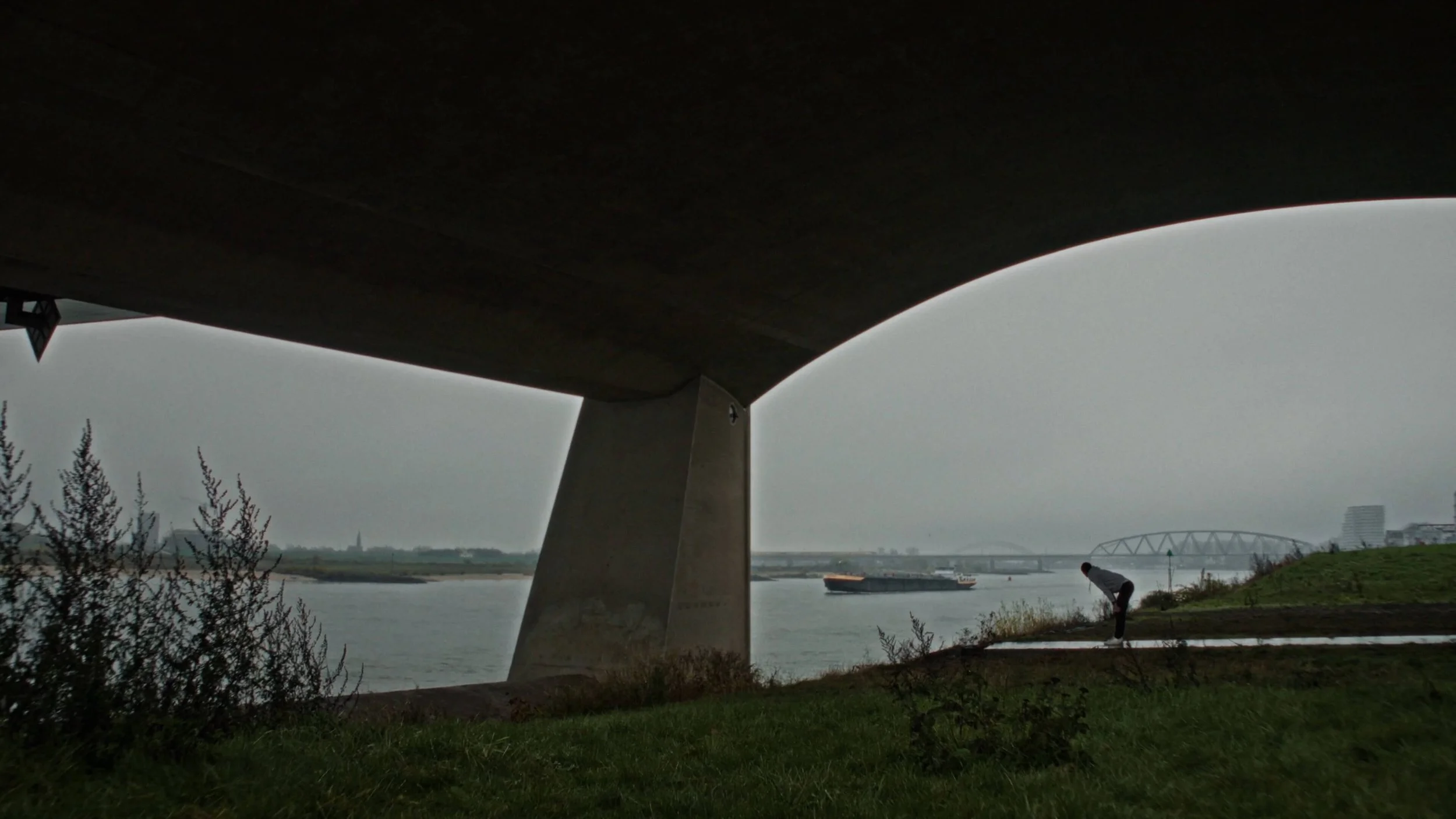 Onder een grote brug staan mensen aan de rivier op een grauwe, bewolkte dag. Er liggen schepen op het water en in de achtergrond is een stad met moderne gebouwen zichtbaar.