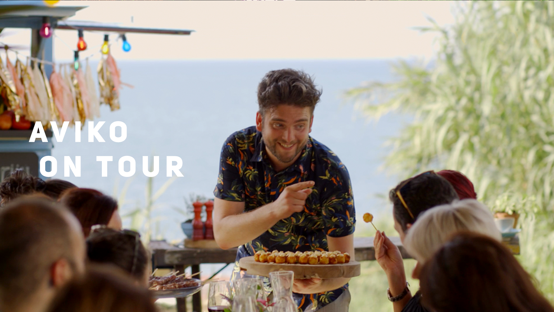 Man serving food at an outdoor event with a sign reading,Table with people and sea view in the background.