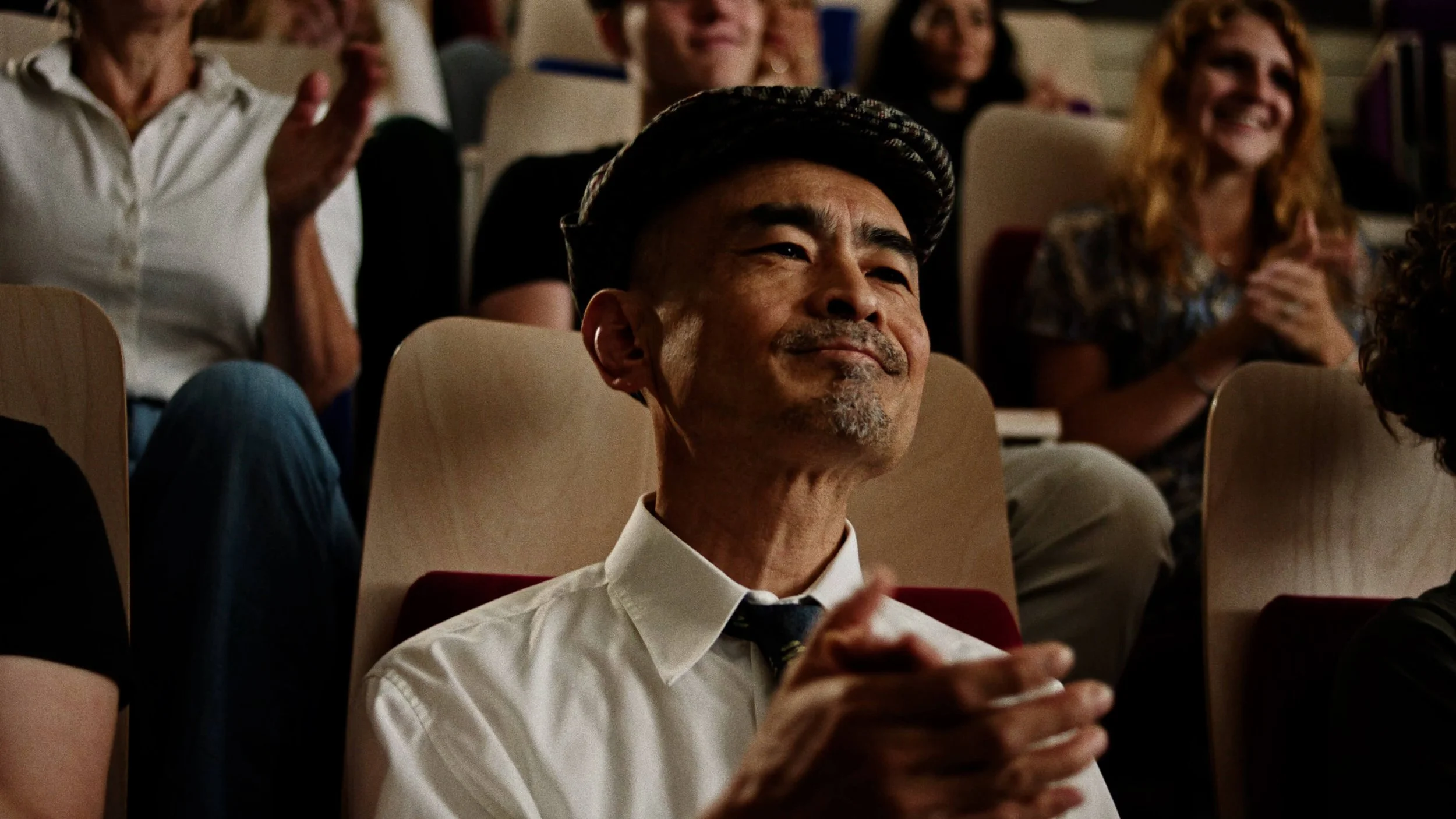 Een man in een witte blouse en een grijze hoed die glimlacht en applaudisseert in een zaal vol lachende mensen.