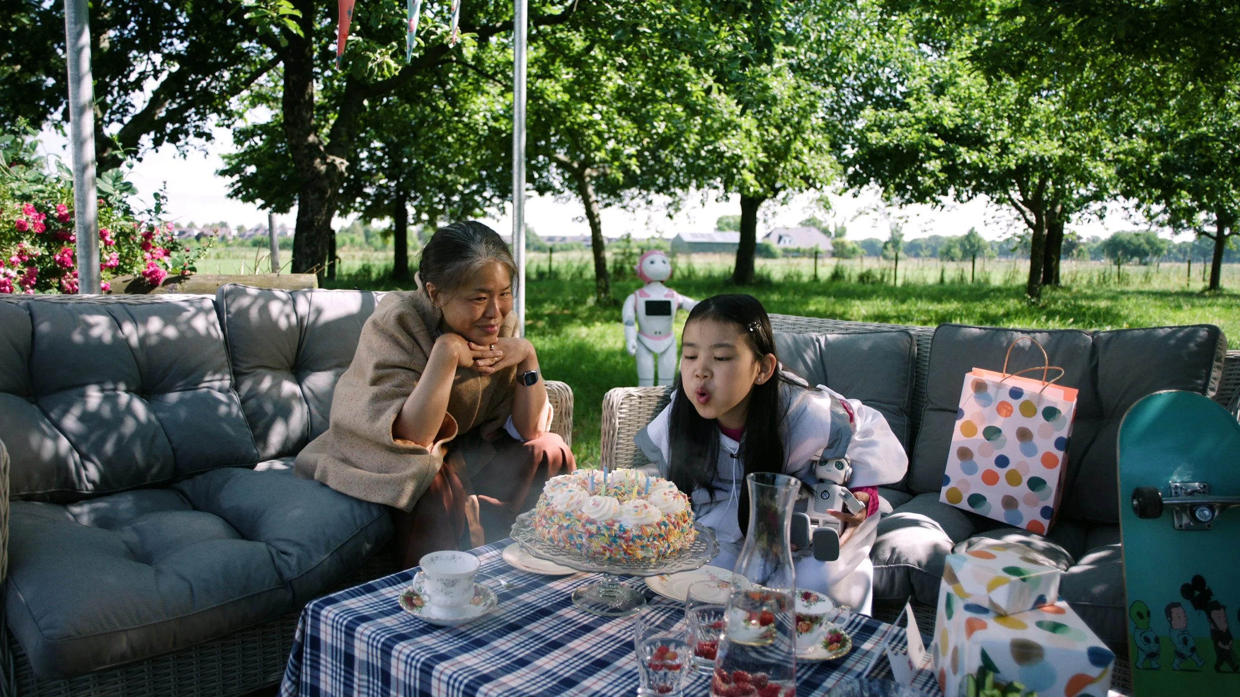 Een jong meisje blaast kaarsen uit op een verjaardagstaart tijdens een buitenfeest met een oudere vrouw die toekijkt, onder een boom met groene bladeren, met cadeaus, drinken en een robot in de achtergrond.