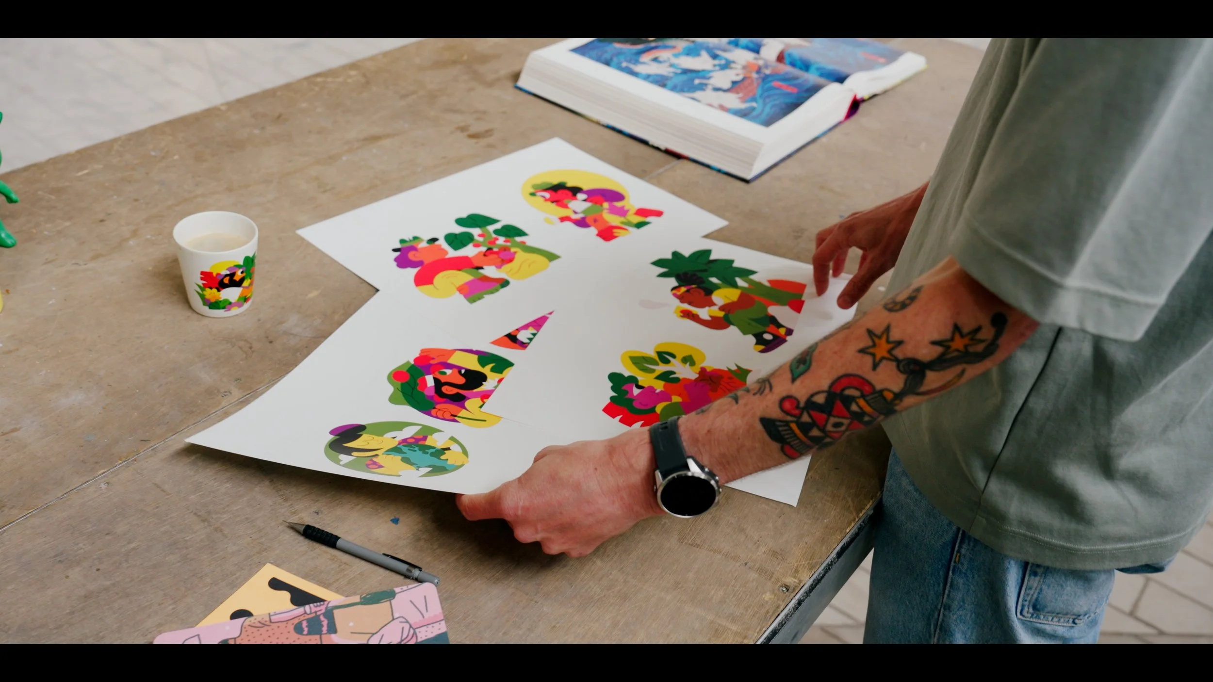 Persoon met tattoos die kleurrijke kunstwerken bestudeert op een tafel met tekeningen en een boek.