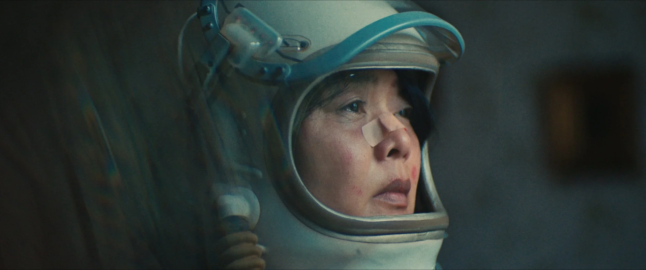 Close-up van een vrouw in een ruimtepak met een pleister op haar neus, met een neutrale uitdrukking, kijkend naar iets buiten beeld.