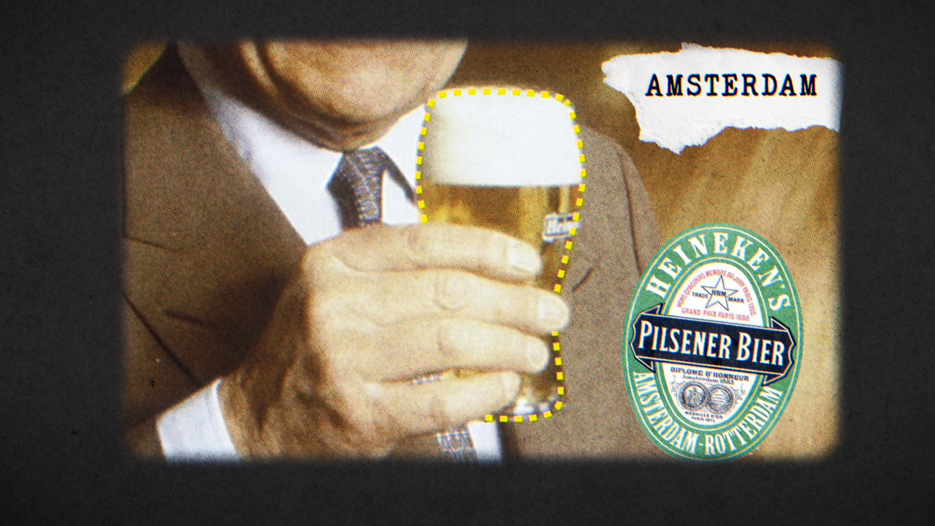 Man in een bruin pak en stropdas houdt een glas pilsener bier, met een etiket van Heineken in de achtergrond.
