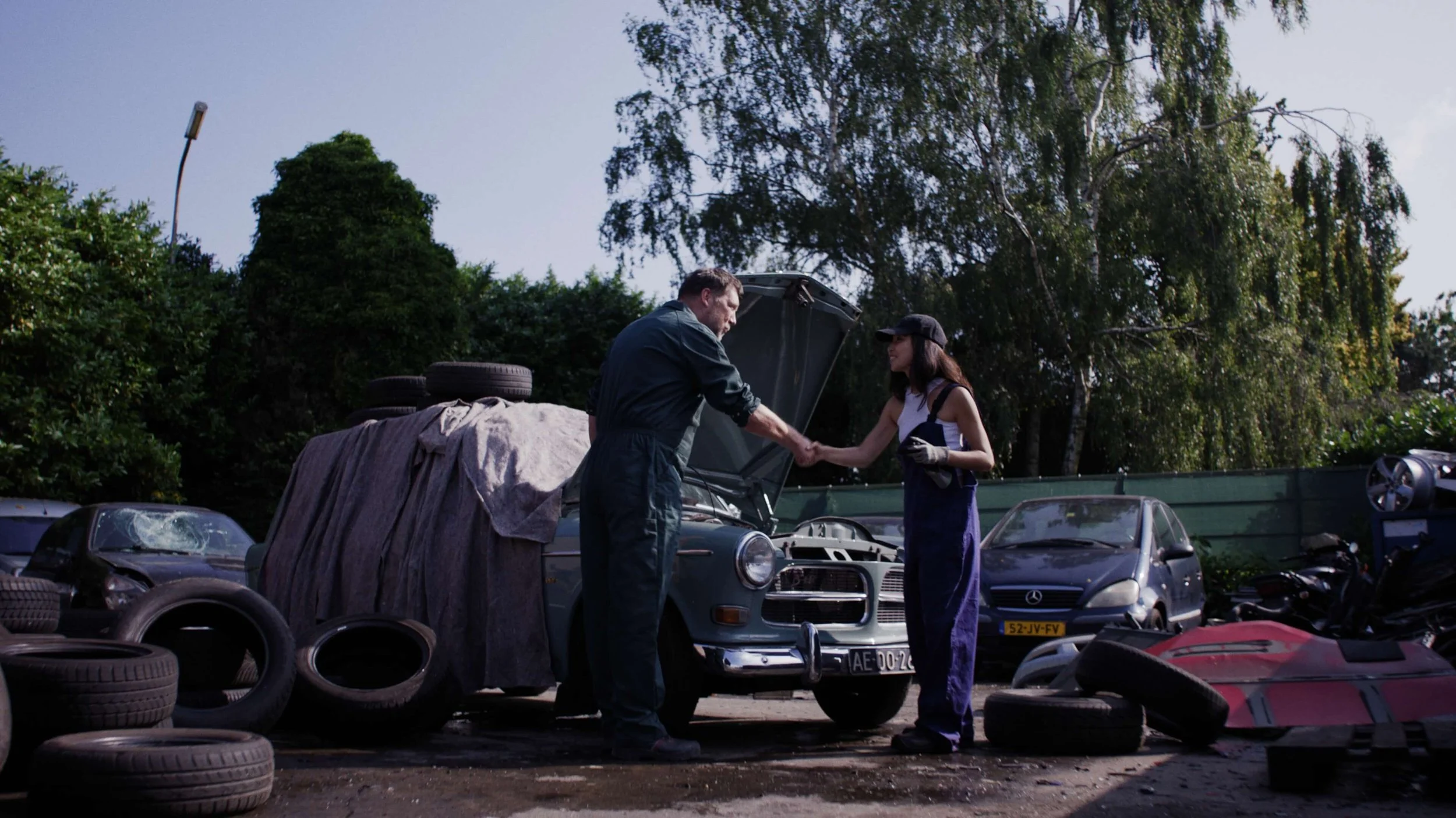 Twee mensen die elkaar de hand schudden op een autosloop, omringd door oude auto's en banden.