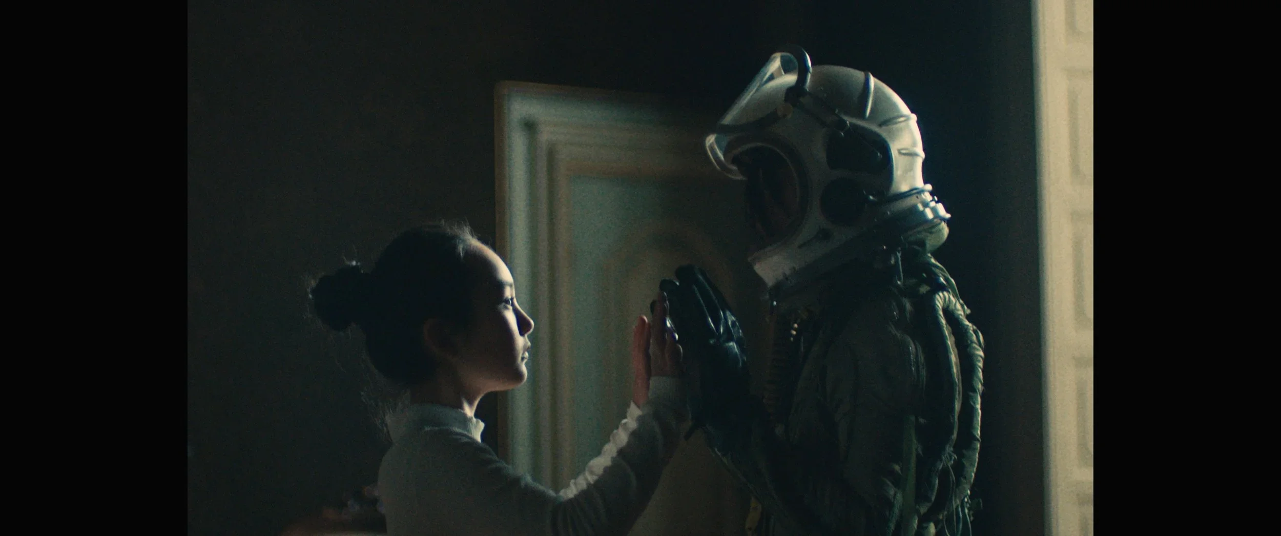 Een jonge vrouw en een astronaut met een ruimtepak en helm die tegen elkaar leunen en met hun handen elkaar aanraken in een dim verlichte kamer.