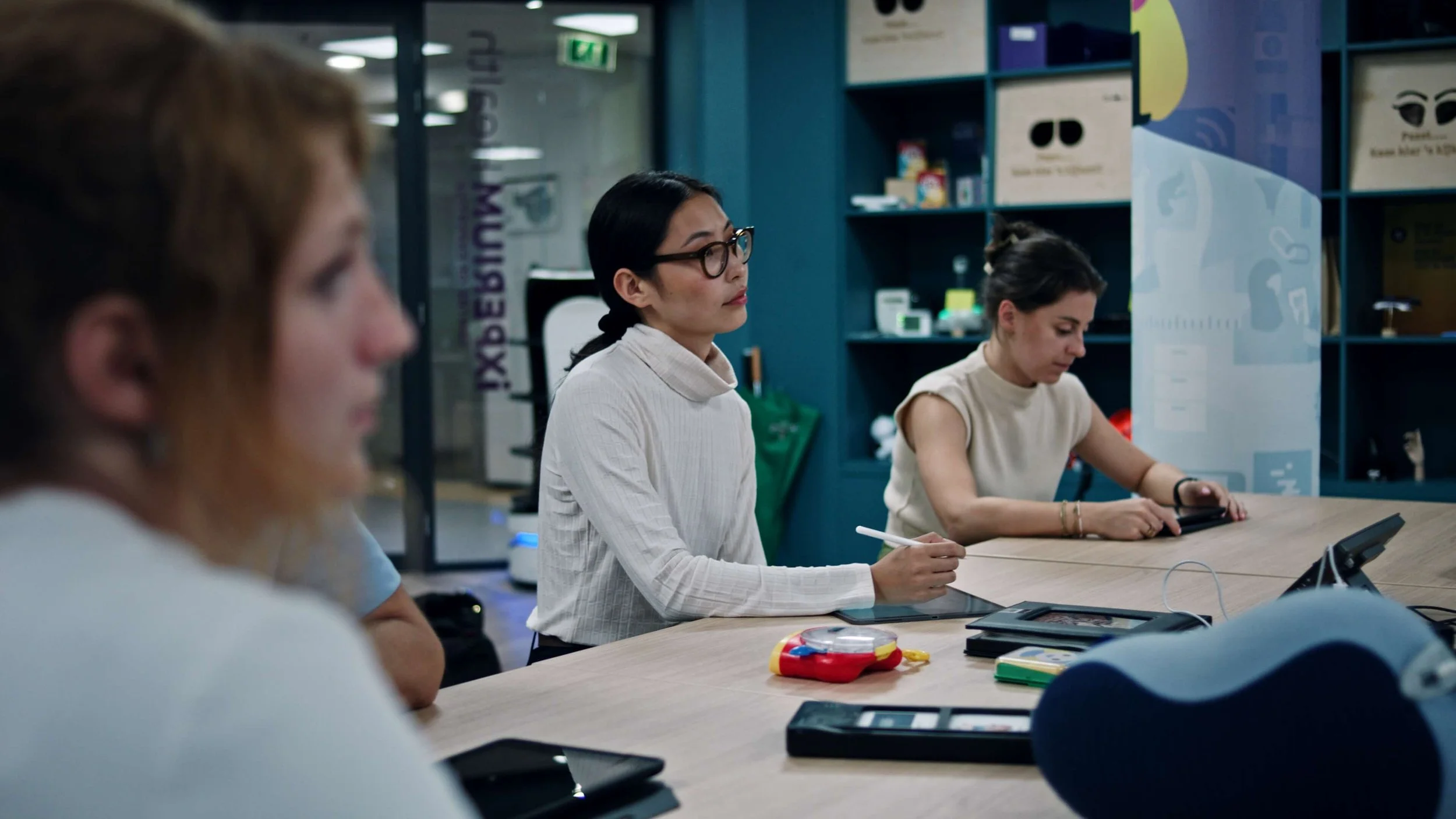 Vier mensen zitten aan een tafel in een moderne vergaderruimte, drie vrouwen en een man, met technologische apparatuur en spelletjes op de tafel.