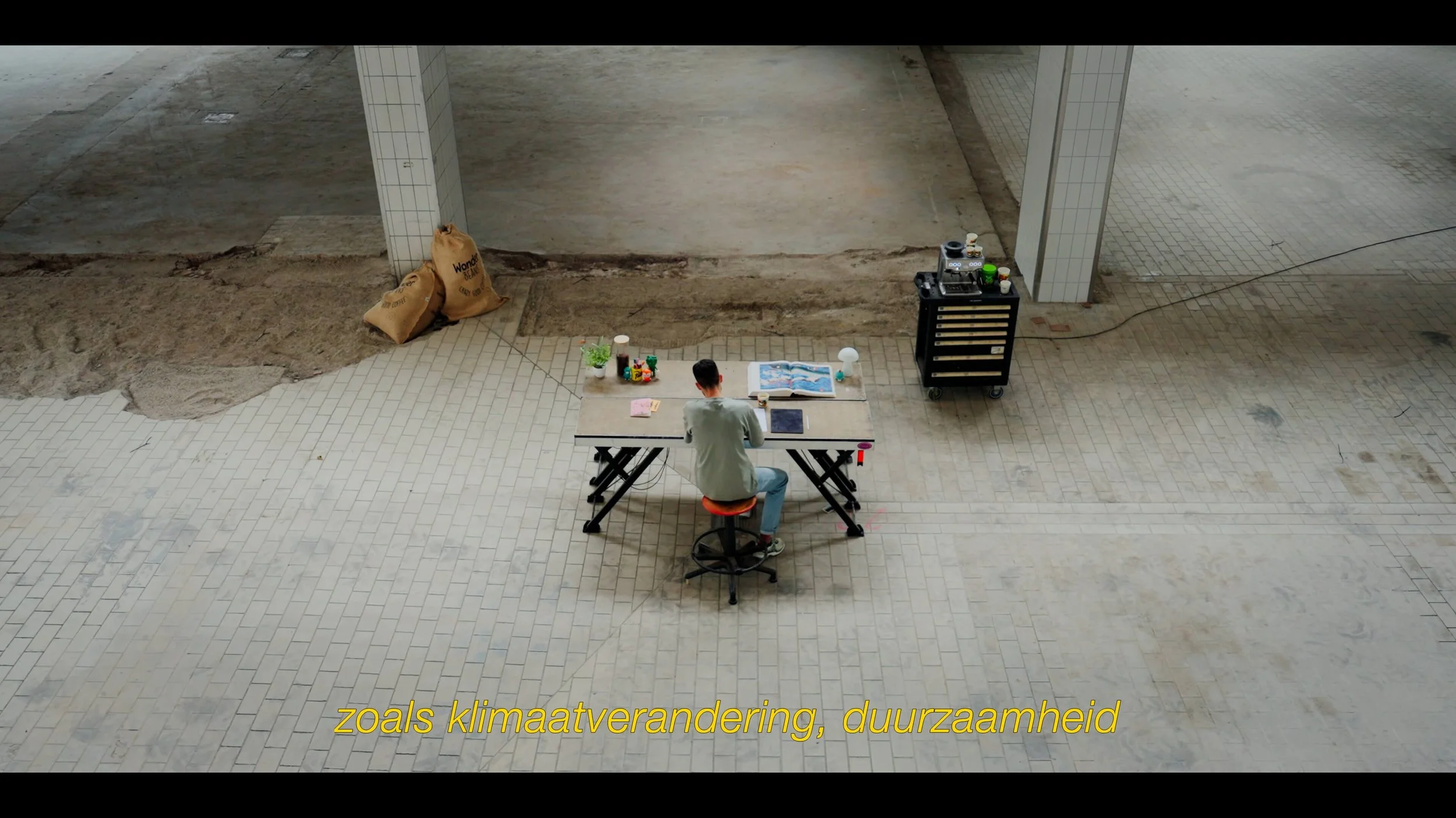 Persoon zit alleen aan een tafel in een grote, open, half afgewerkte ruimte met bakstenen vloer. Op de tafel liggen een open boek, plantjes, een tablet en kantoorbenodigdheden. Aan de rechterkant staat een op rolletjes kast met apparatuur.