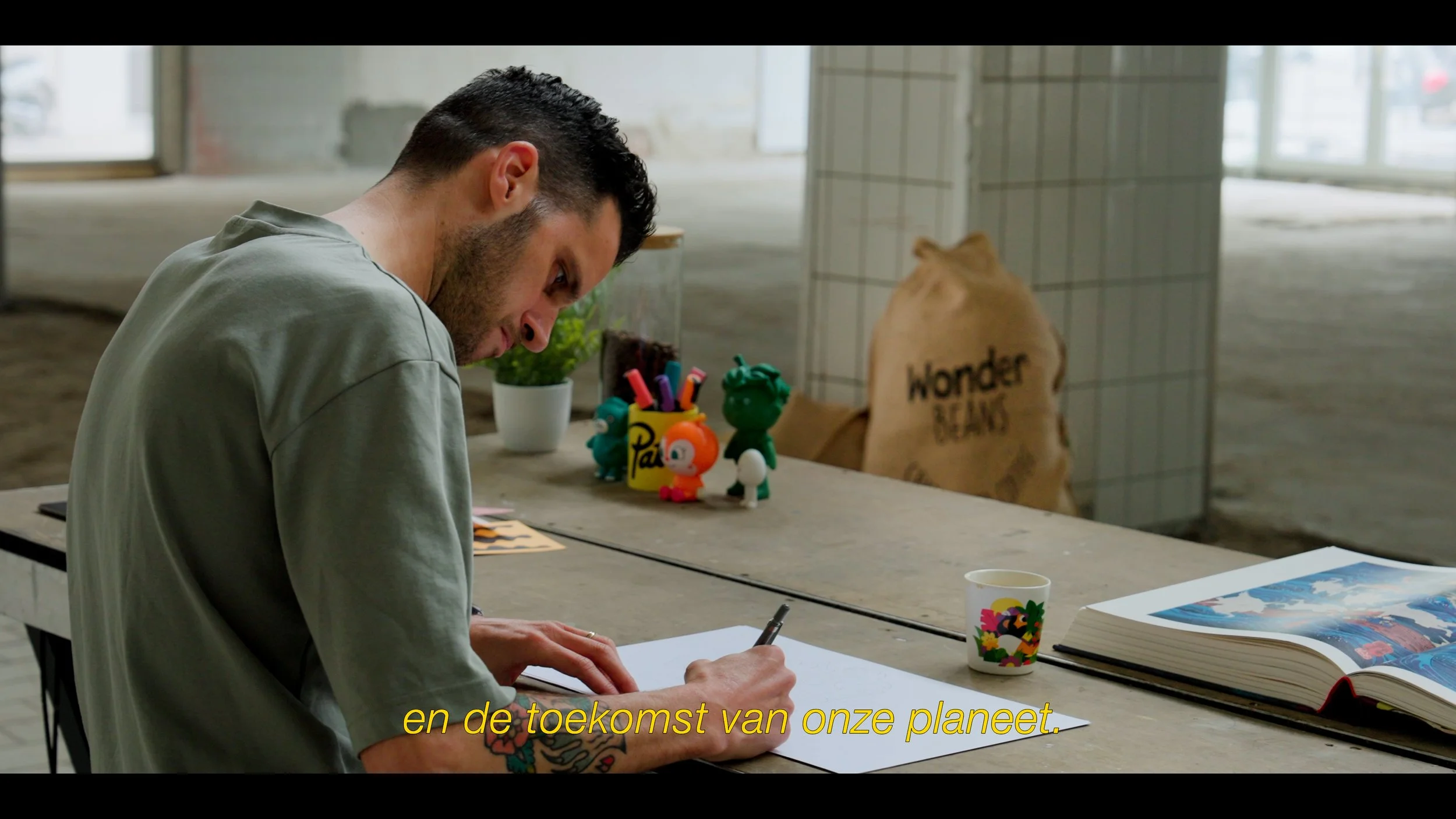 Man zit aan een tafel, schrijft op papier, met kleurrijke figurines, een beker en een open boek op de tafel, in een industrieel ogende ruimte.