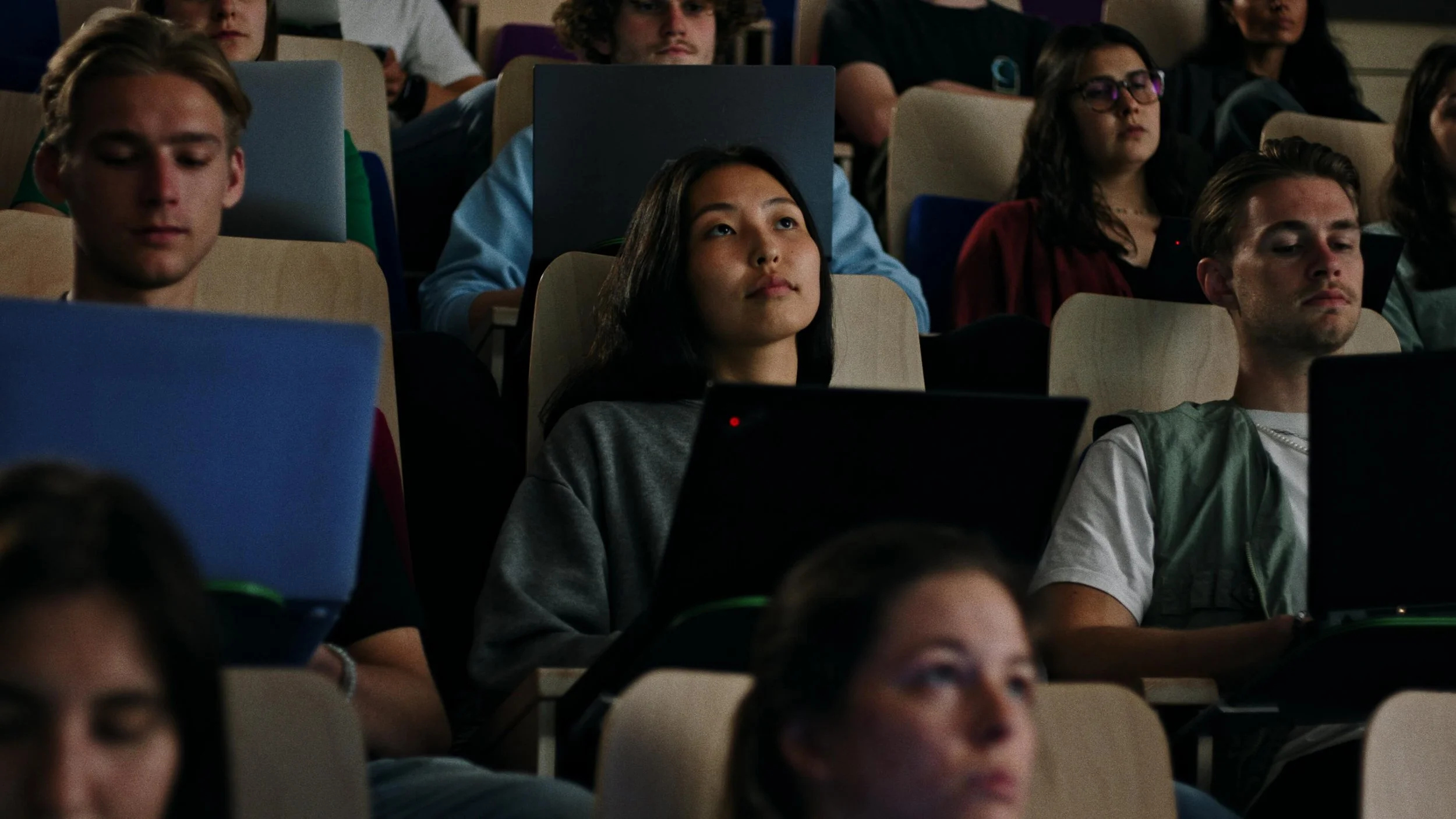 Een groep jonge studenten zit in een collegezaal, met laptops open voor hen, en ze lijken geconcentreerd te luisteren of te werken.