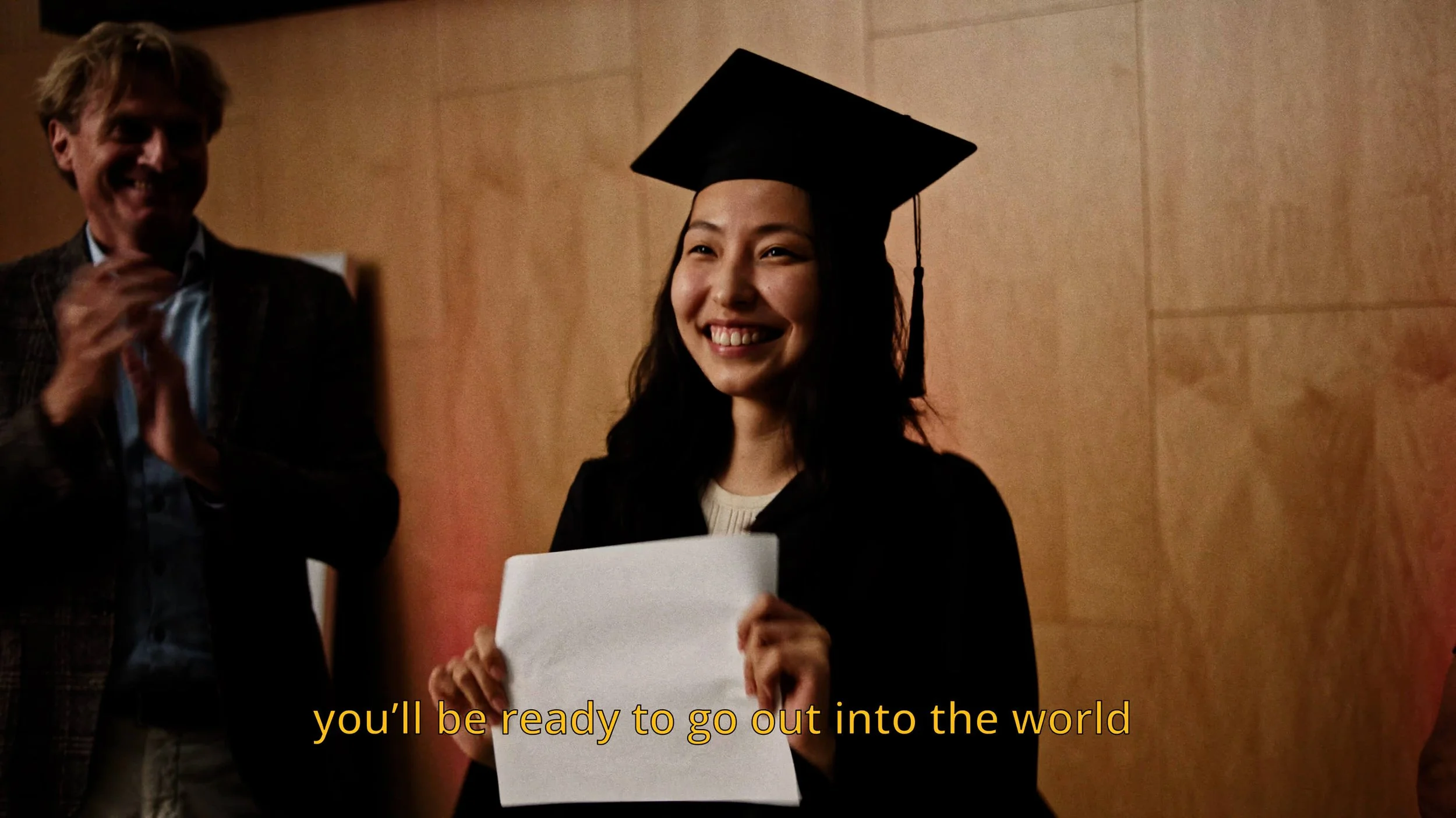 Een jonge vrouw met een afstudeerhoedje die een brief vasthoudt, glimlachend tijdens haar afstudeerceremonie, met een man die applaudisseert op de achtergrond.