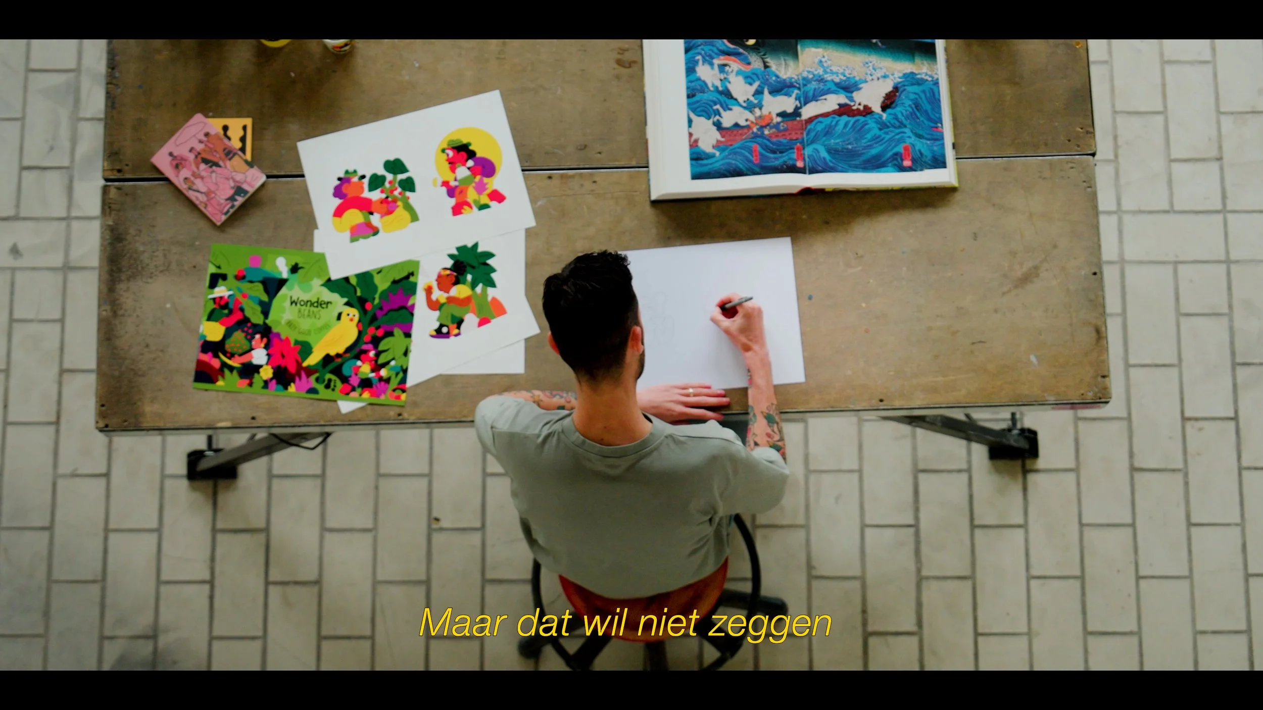 Man zit aan een houten tafel en tekent op een stuk papier. Op tafel liggen kleurrijke kunstwerken, waaronder platte illustraties van mensen en een afbeelding met een papegaai, en een open boek met een Japanse kunstprint. Er is een subtitel onderin de