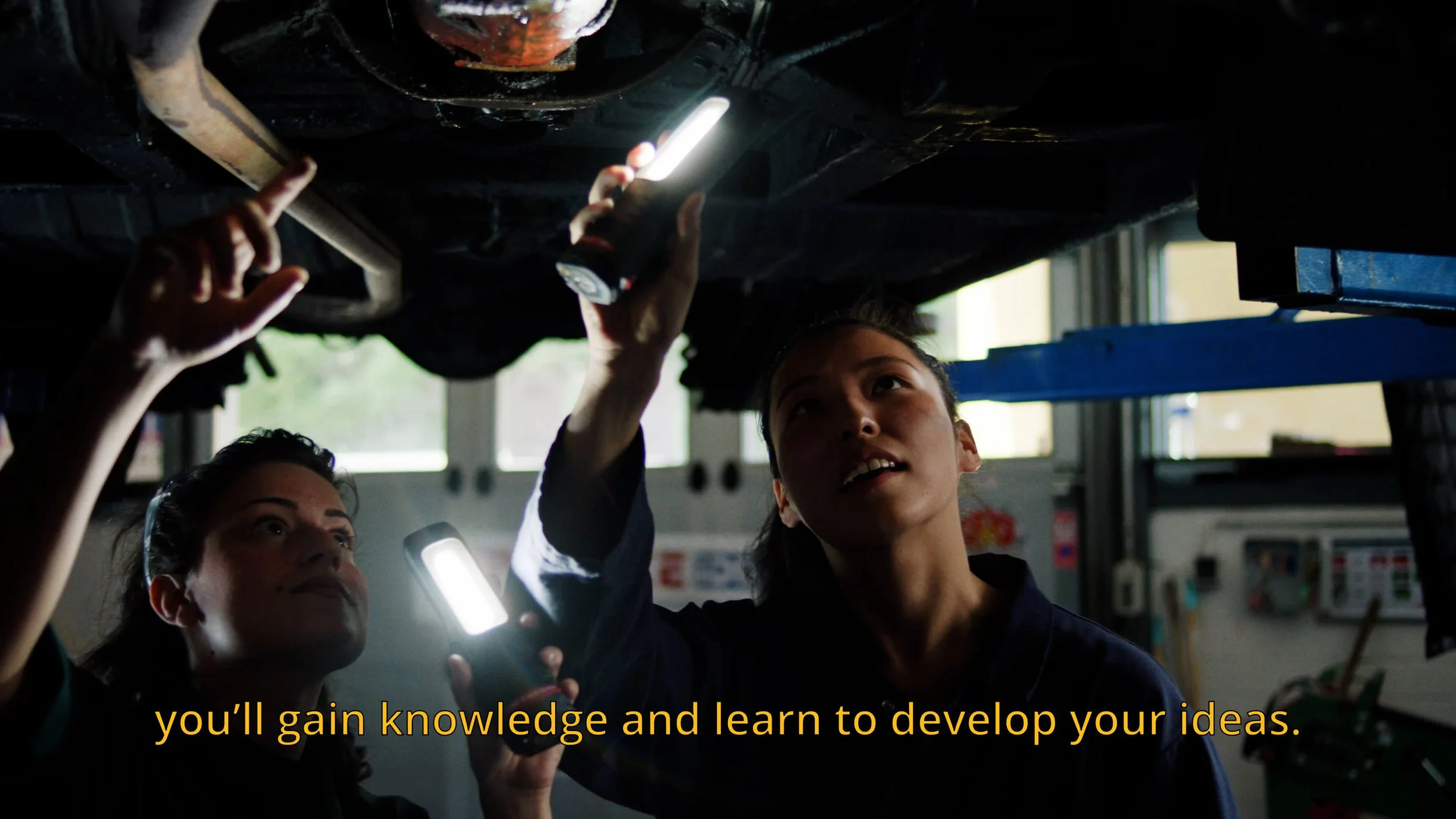 Twee vrouwen inspecteren de onderkant van een auto in een workshop, een van hen houdt een zaklamp en wijst naar de auto, terwijl de andere naar de auto kijkt.
