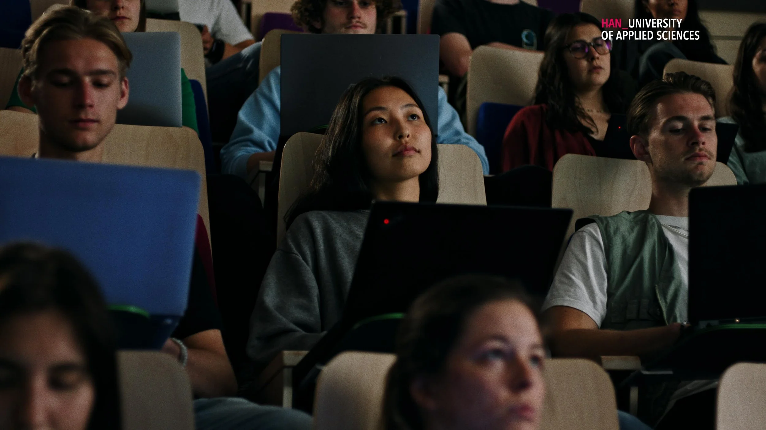 Studenten zitten in een collegezaal met laptops, sommigen hebben hun ogen gesloten of kijken nadenkend.