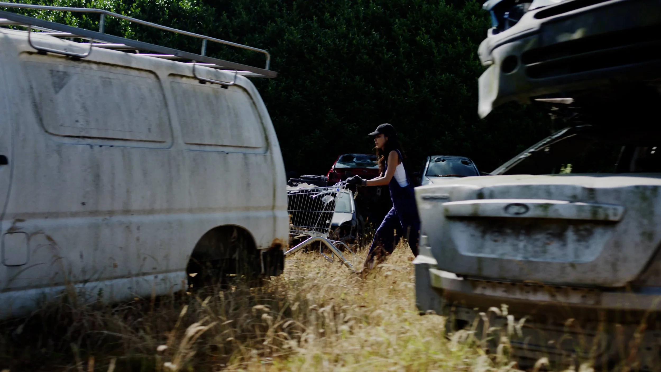 Een vrouw met een pet duwt een winkelwagen tussen verwaarloosde, oude voertuigen in een grasveld.