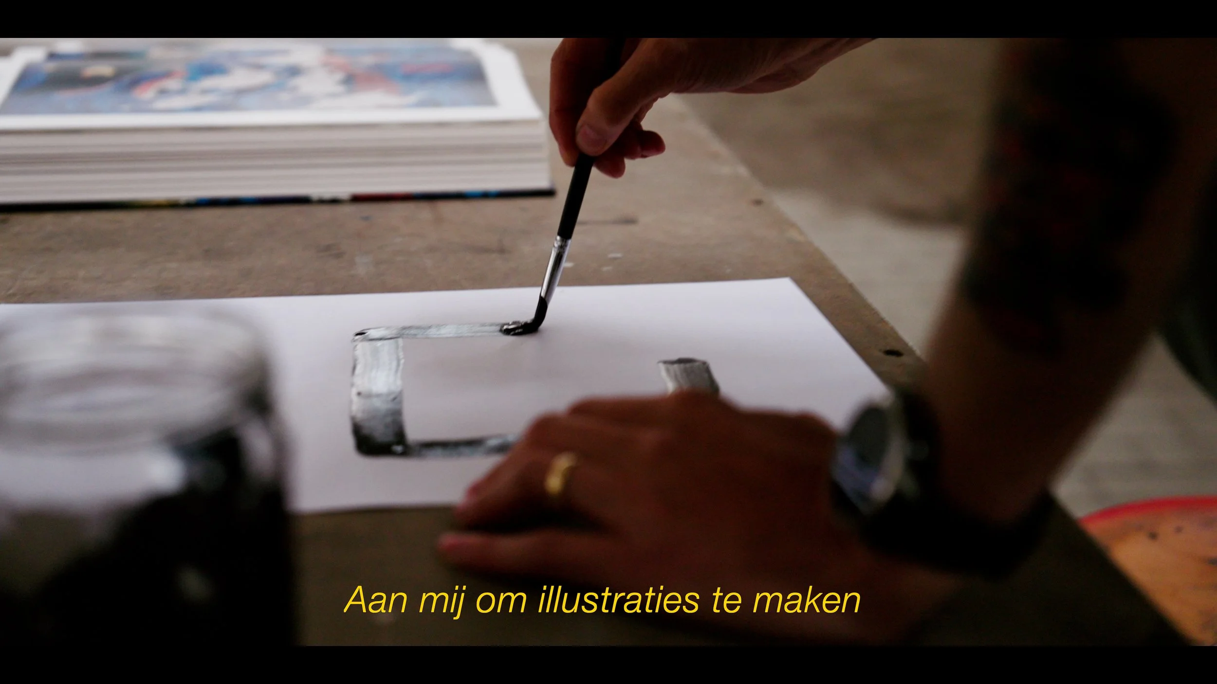 Persoon die met een penseel op een stuk papier tekent, met een comicboek of tijdschrift op de achtergrond en een pot verf op de tafel, met Nederlandse ondertitel 'Aan mij om illustraties te maken'.