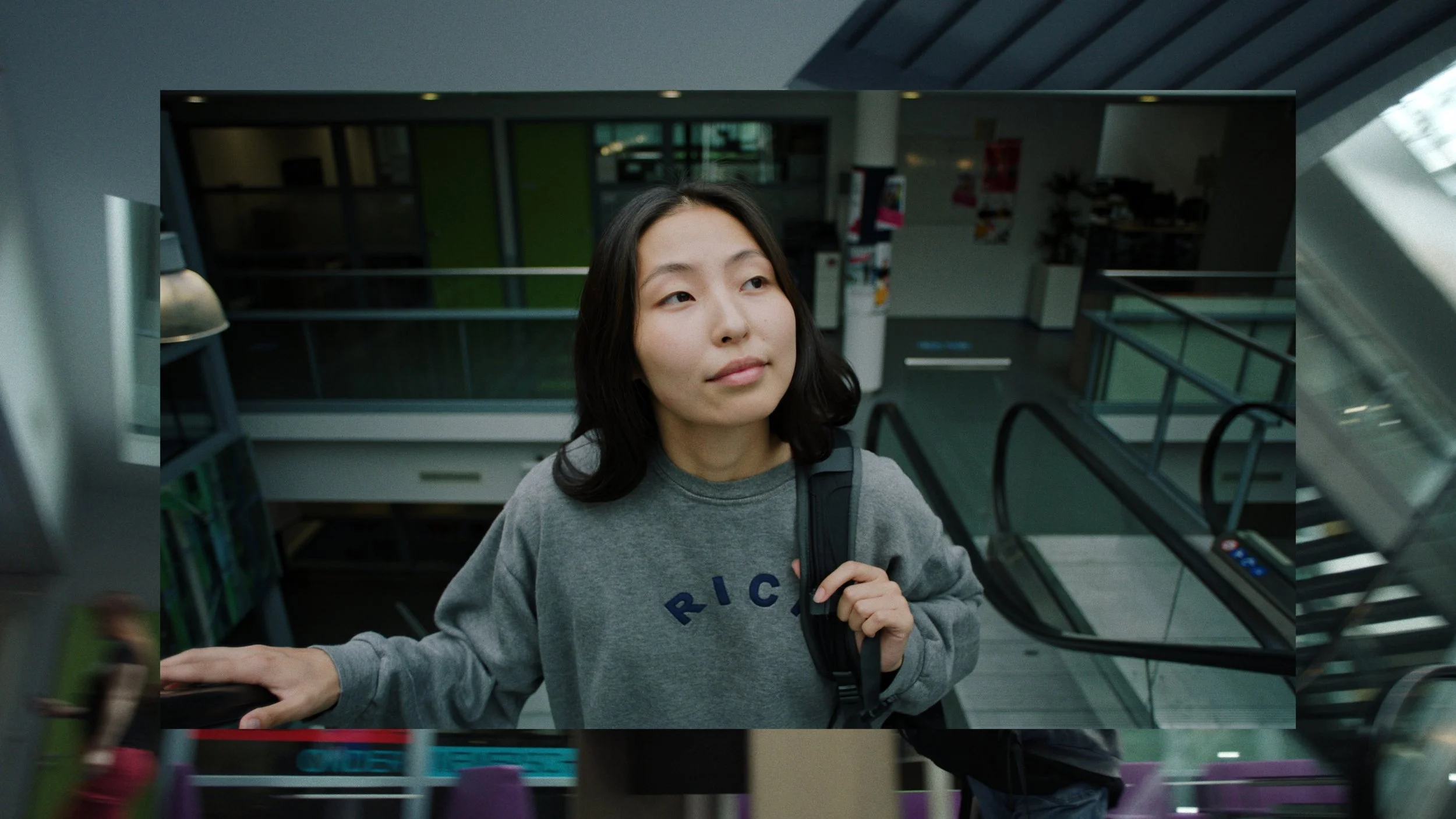 Een vrouw met zwart haar in een grijze trui die op een roltrap staat, met een backpack over haar schouder in een gebouw met grote ramen en groene muren.