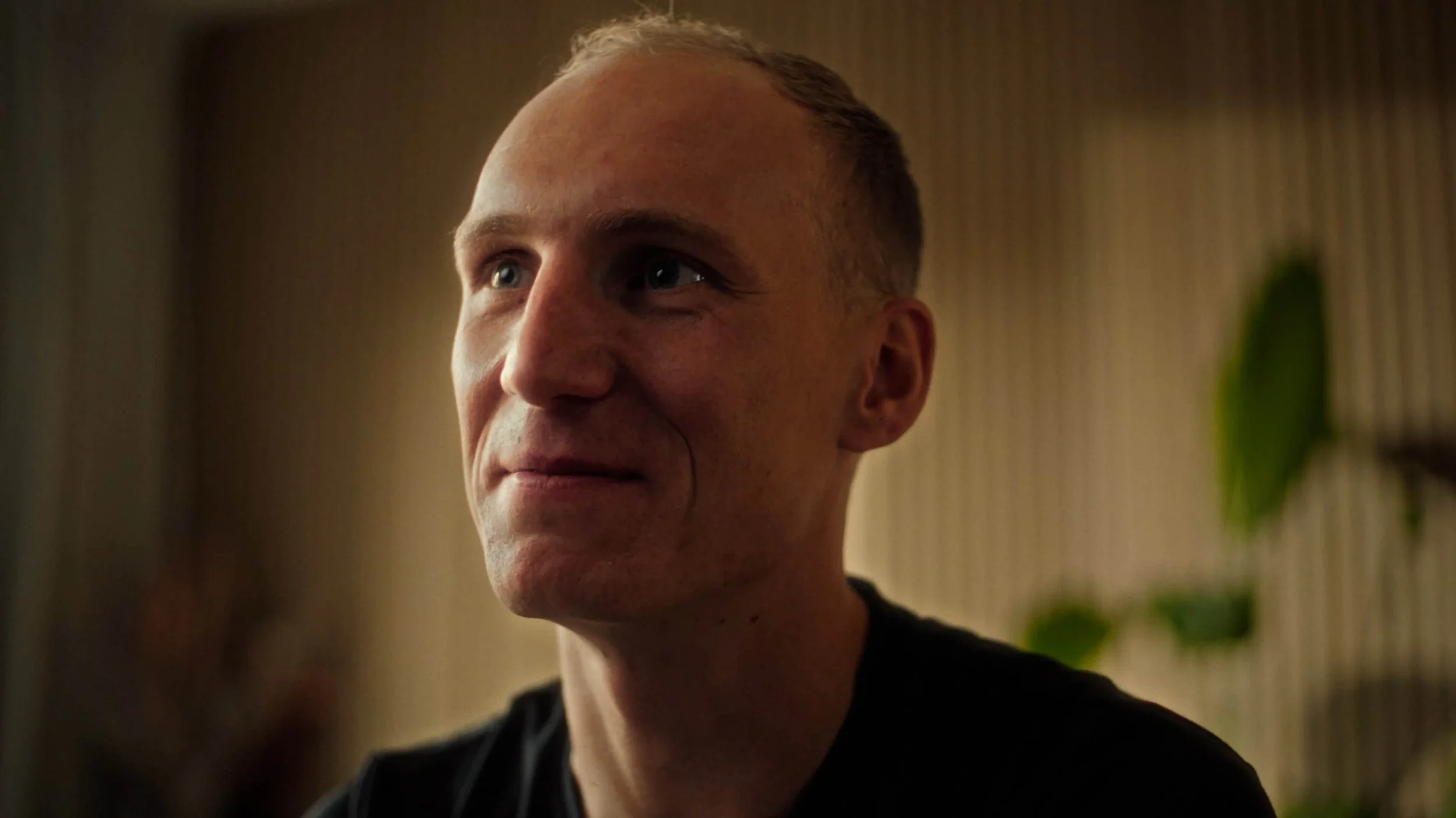 Portret van een man met een korte haarstijl, in een binnenruimte met een patroon op de muur en wat groen blad op de achtergrond.