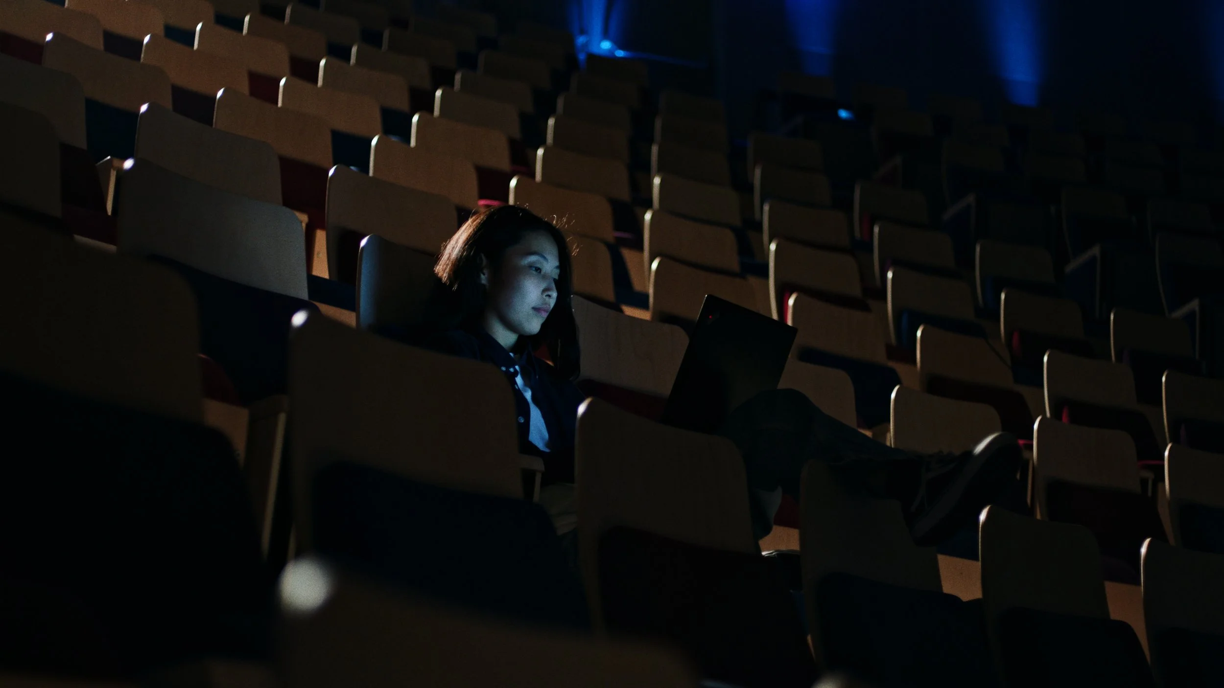 Een vrouw zit alleen in een grote, donkere zaal met lege houten stoelen en kijkt naar een laptop die haar in het gezicht verlicht.