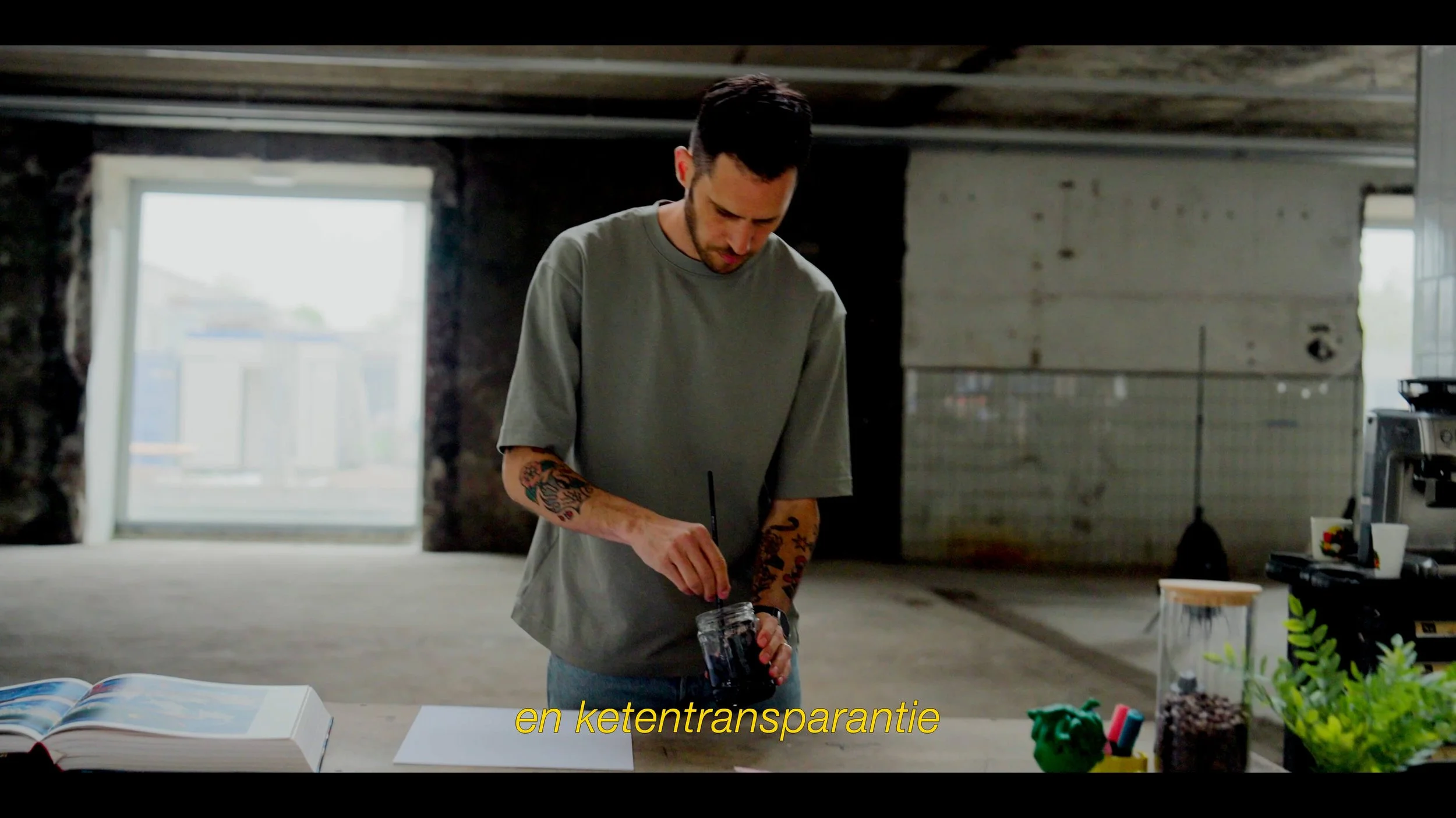 Man die in een open ruimte suikerstroop of een soortgelijke substantie in een pot roert, met planten en andere entiteiten op tafel en grote ramen op de achtergrond.