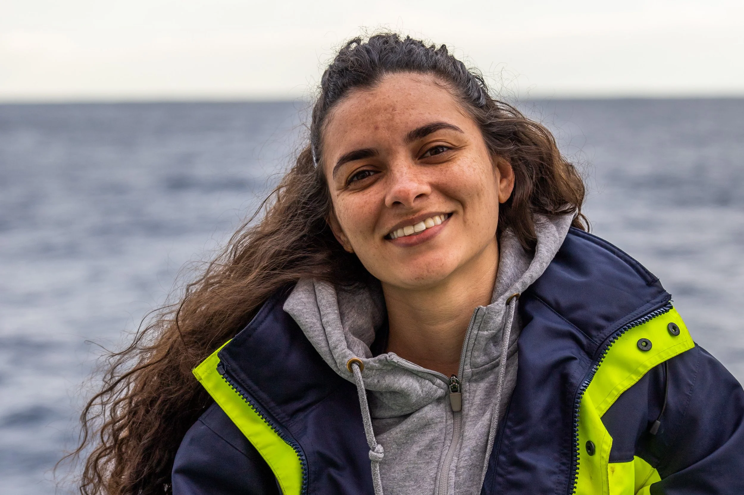 A young woman with long, wavy brown hair smiling outdoors by the sea, wearing a navy and neon yellow jacket over a gray hoodie.