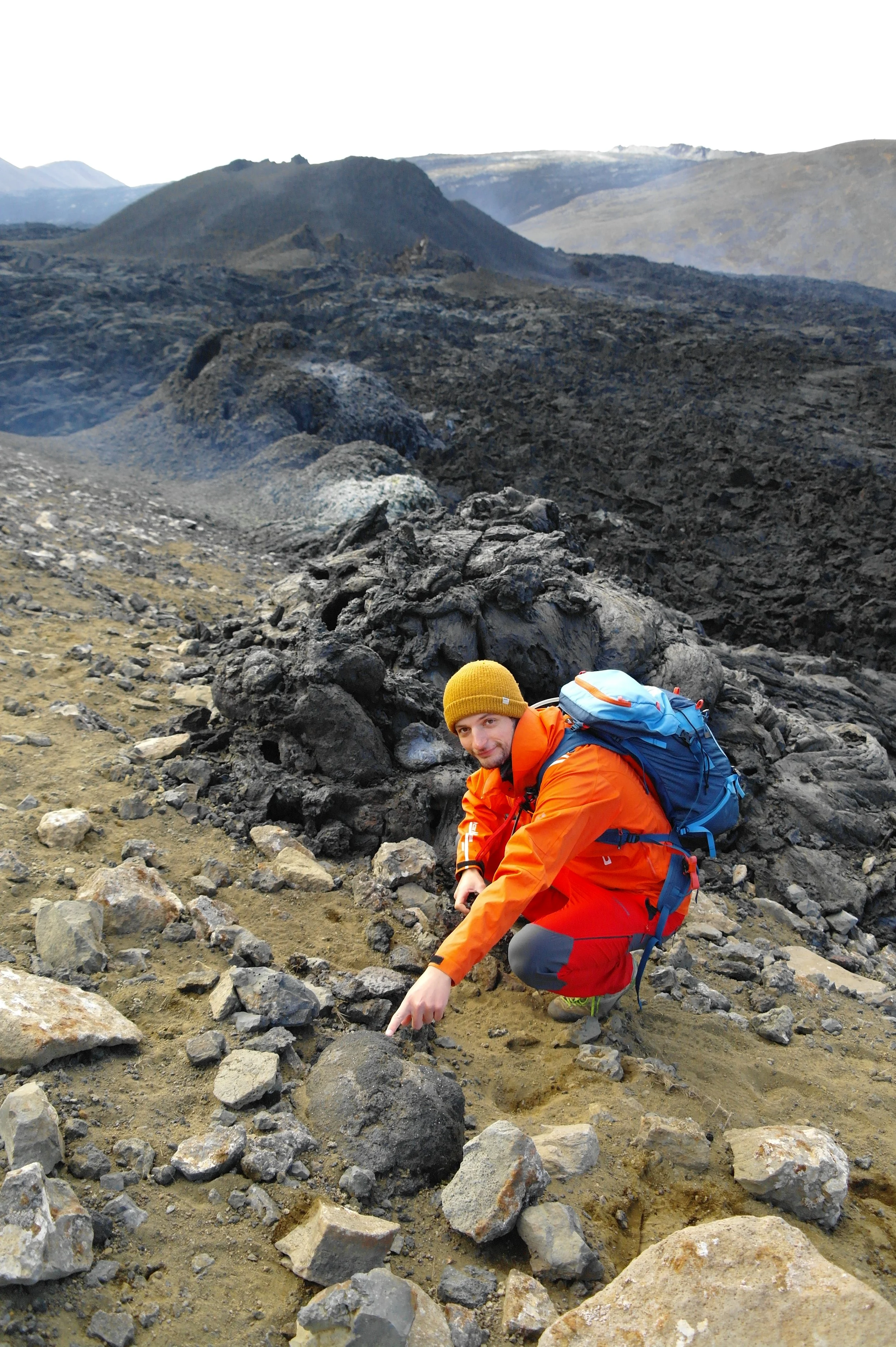 A man in an orange jacket and yellow beanie crouching on rocky terrain with volcanic landscape in the background.