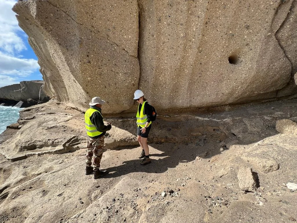 Two people wearing safety vests and white helmets standing on a rocky beach near a large granite cliff.