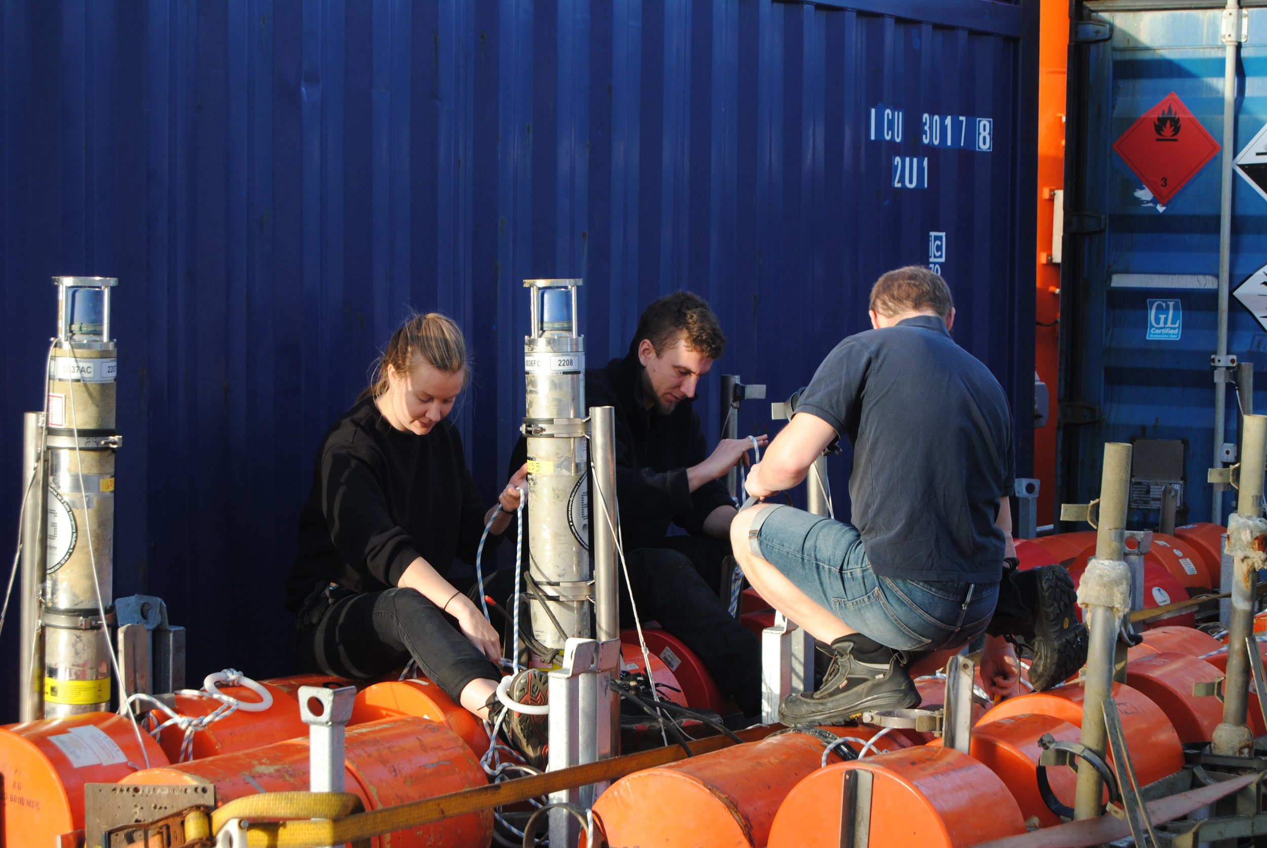 Three people working on fuel tanks in an industrial setting with a blue shipping container in the background.