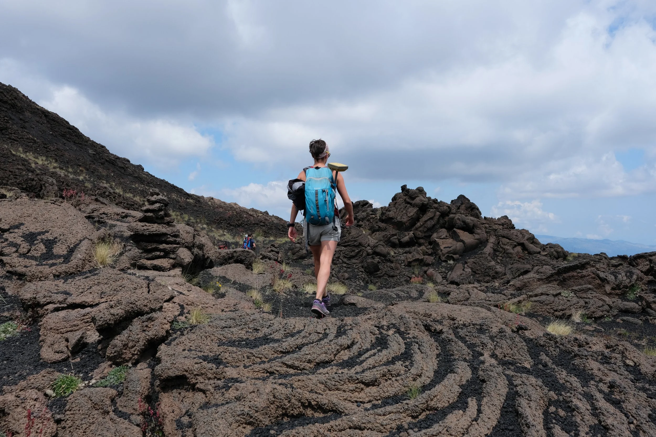 A person hiking on a rocky terrain with a blue backpack and walking sticks, leading with other hikers in the distance under a cloudy sky.